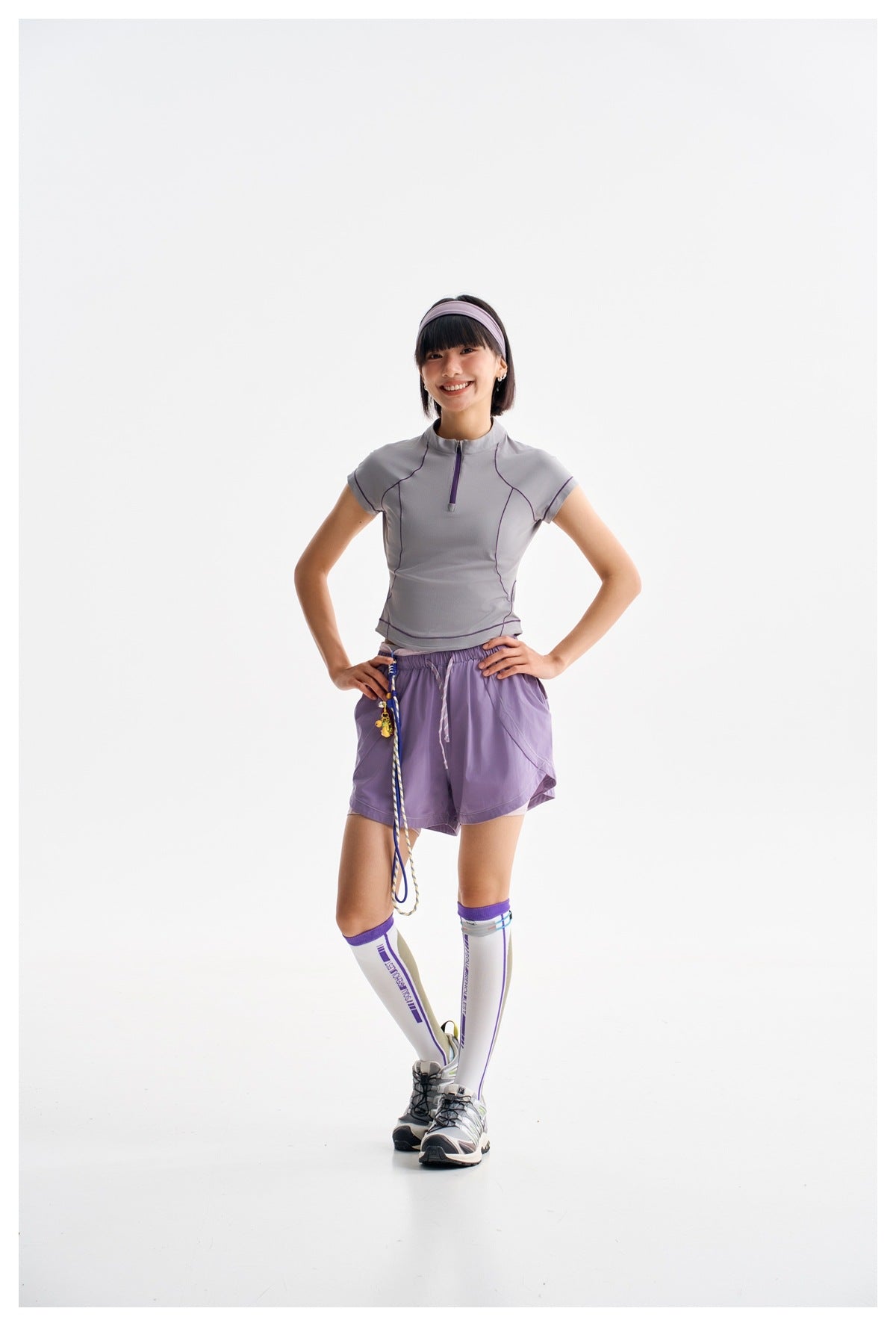 Model wearing a gray-lavender zip-neck top and purple shorts with white knee-high socks and sneakers.