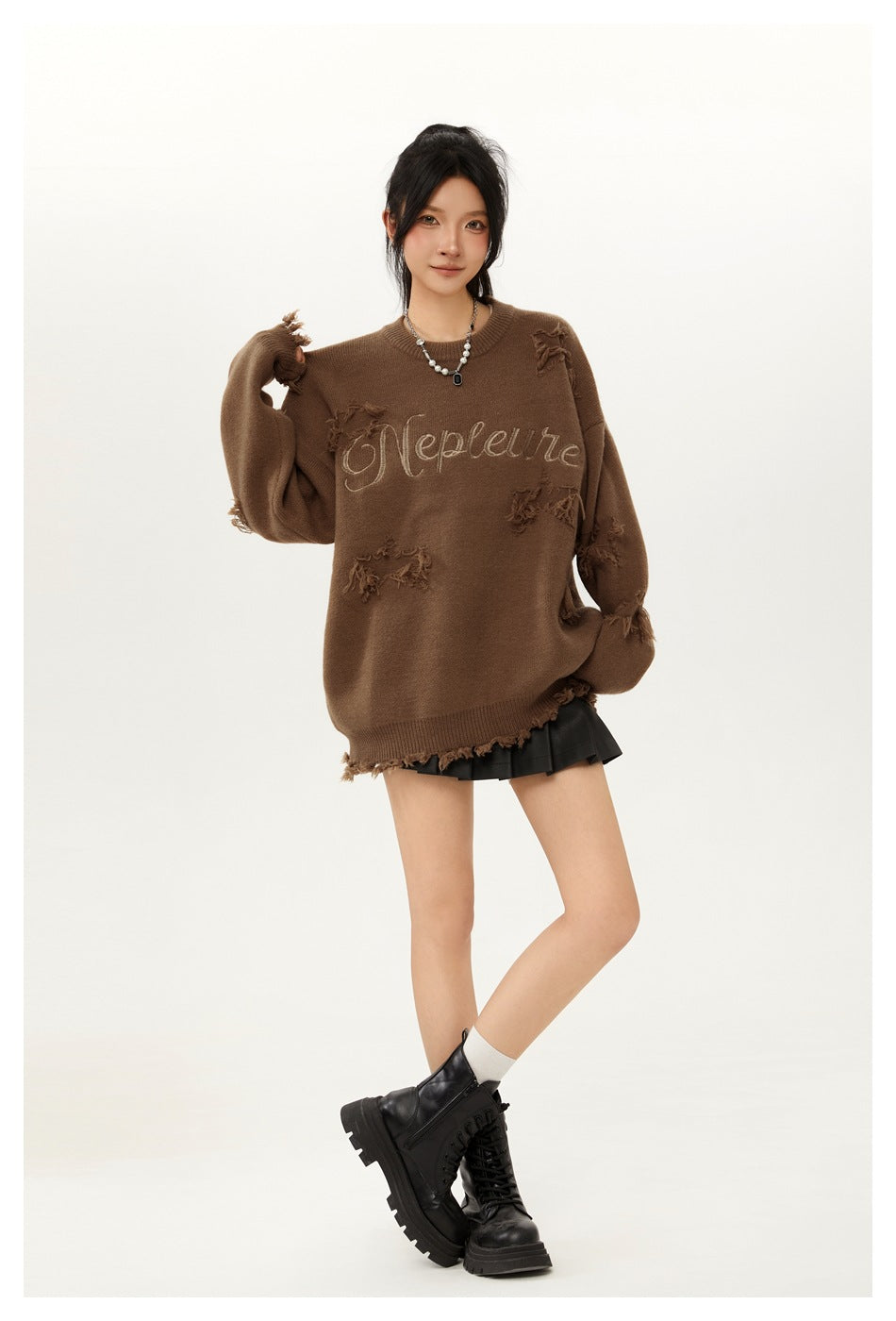Model wearing an oversized brown distressed crewneck sweater with embroidered script and a black pleated mini skirt.