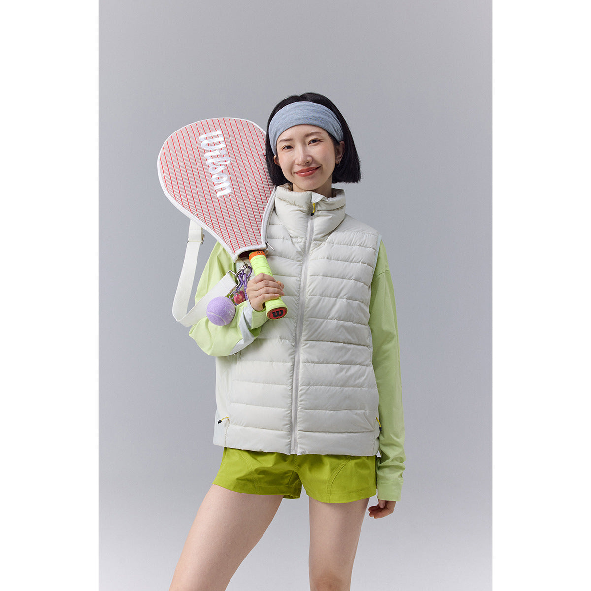 Model wearing a white quilted puffer vest over a lime-green top and shorts, holding a tennis racket.