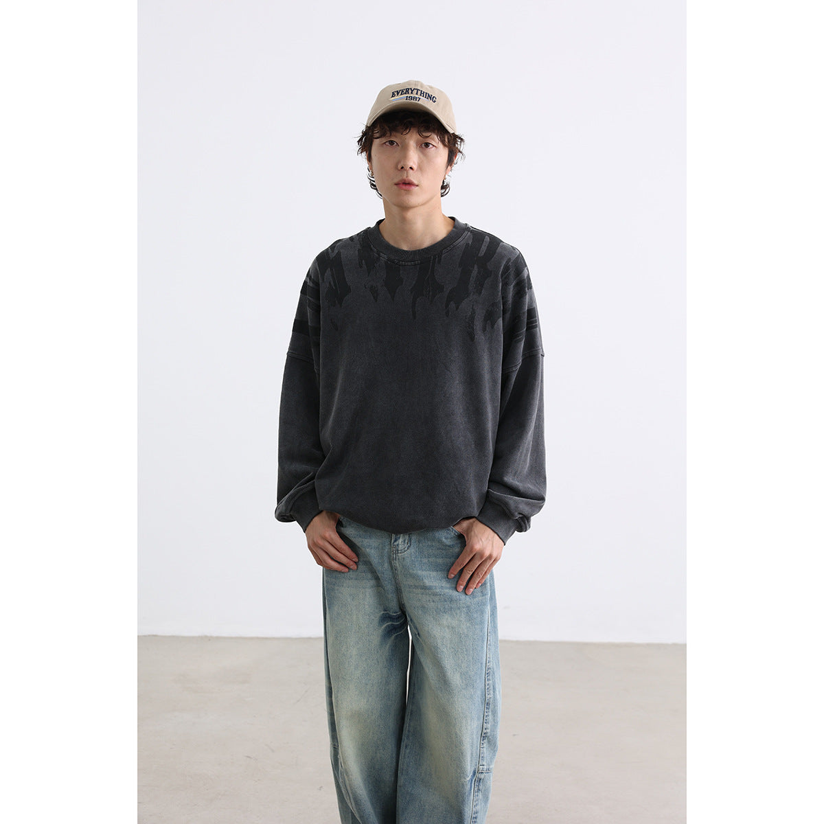 Model wearing a charcoal oversized sweatshirt with dark shoulder graphics, faded blue jeans and a beige cap.
