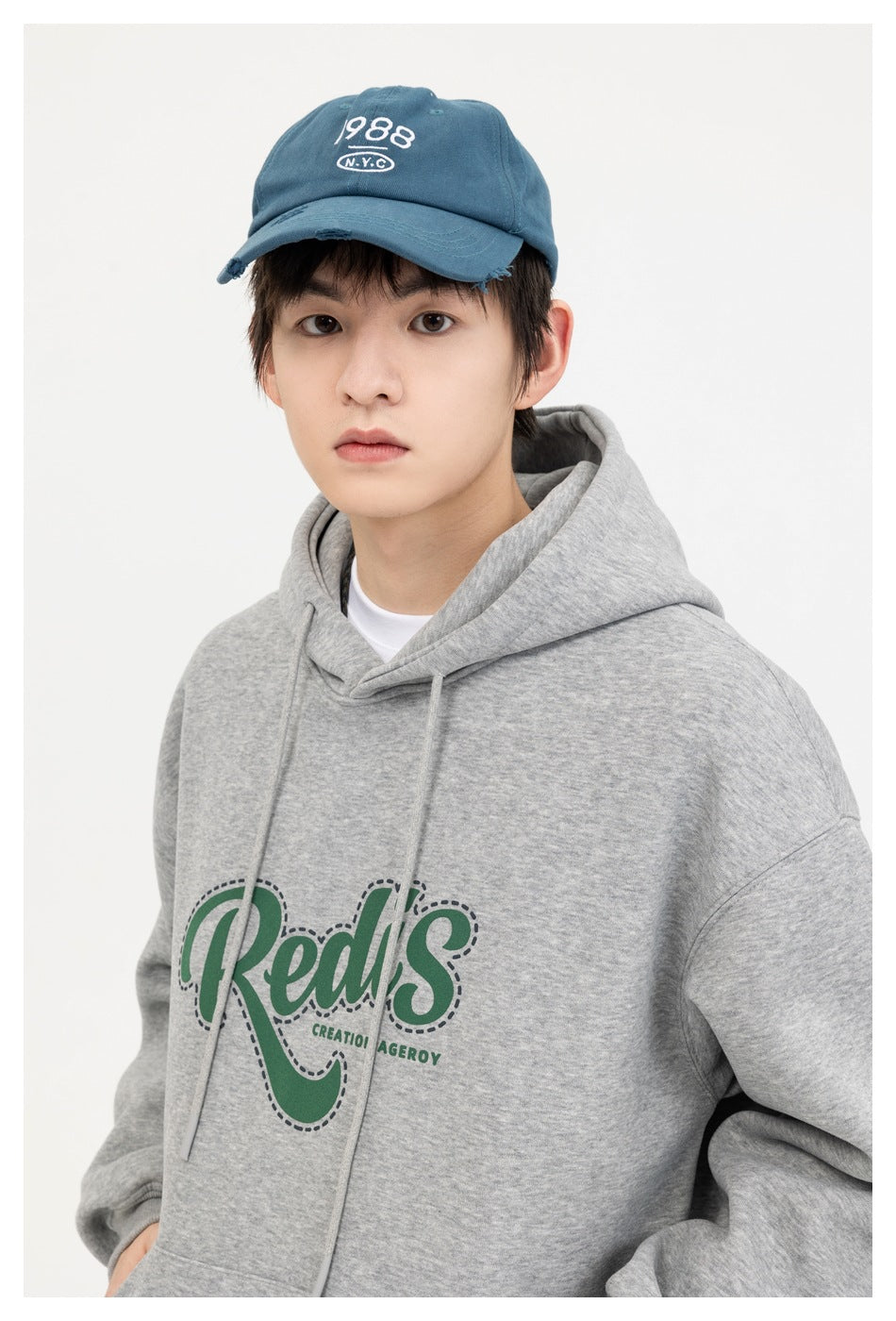 Close view of a model wearing a gray hoodie with green chest lettering and a blue embroidered cap.
