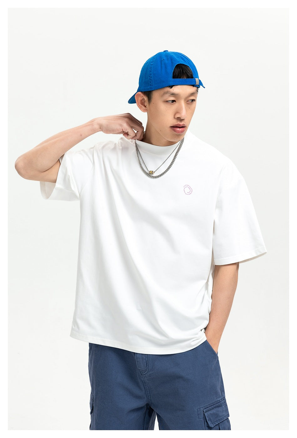 Man wearing an oversized white T-shirt with a small pink circular chest print, blue cap, and blue cargo shorts.