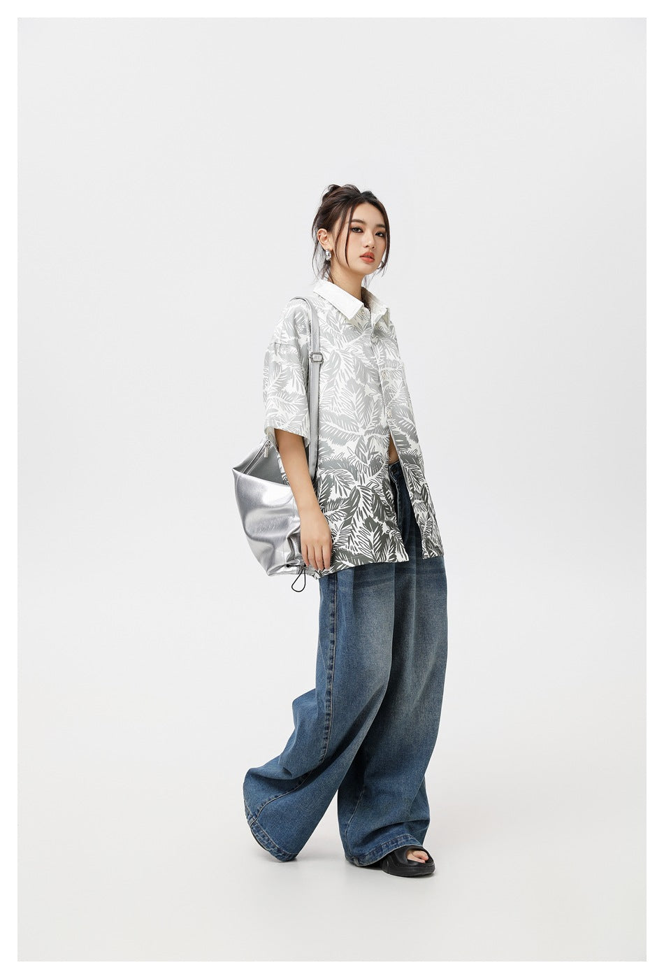 Model in a gray-and-white tropical leaf print shirt and wide blue jeans, shown at an angle with a silver shoulder bag.