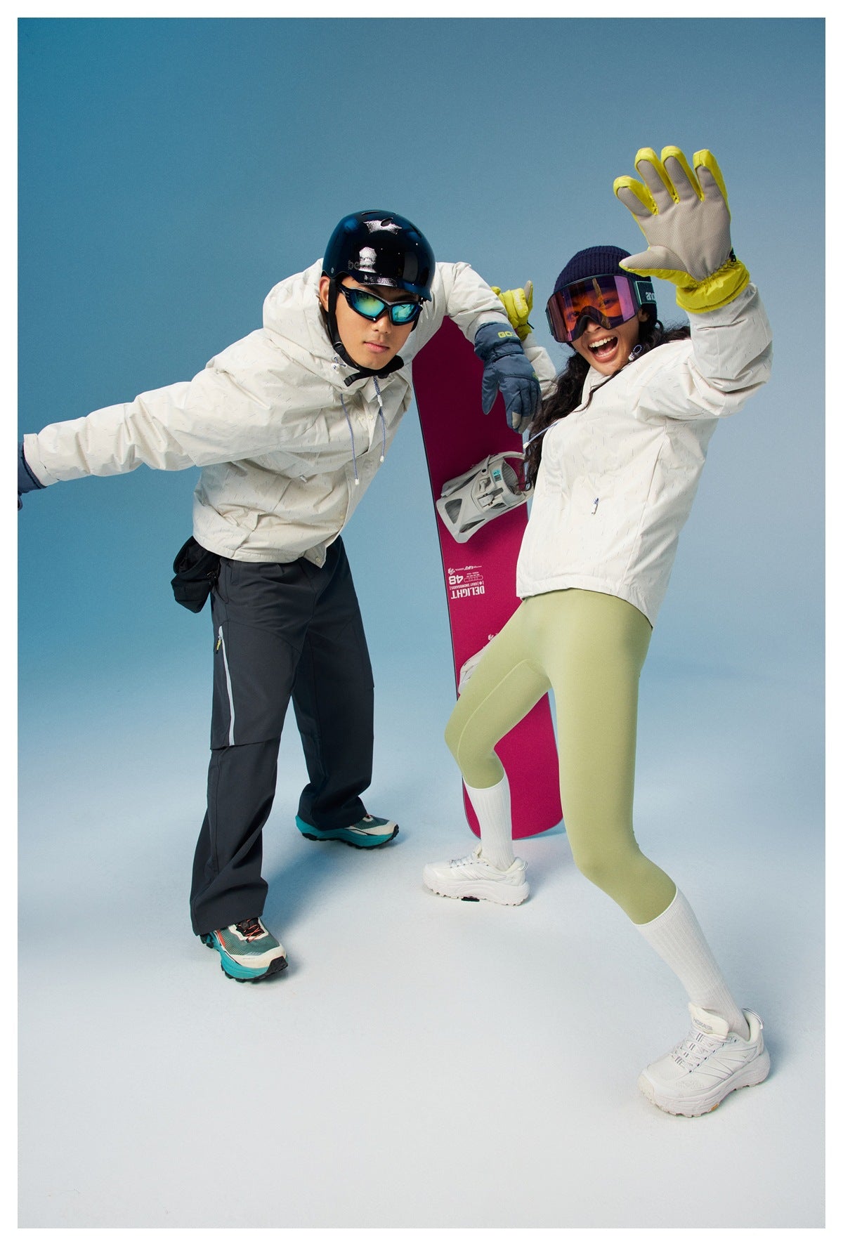 Two snowboarders pose in white jackets; one wears dark pants and the other light green leggings.