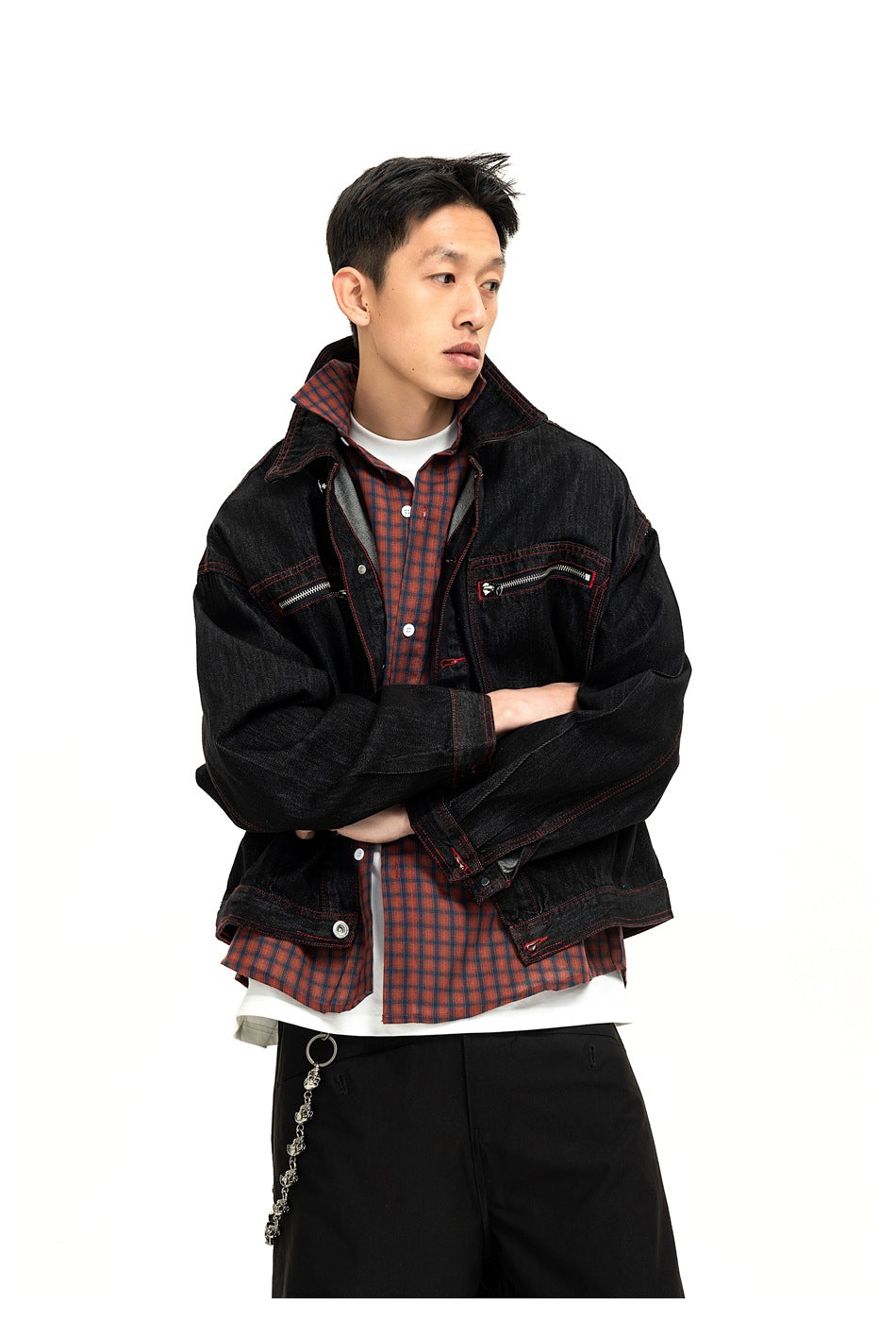 Man wearing a black denim jacket with red stitching over a red plaid shirt, arms crossed, looking to the side