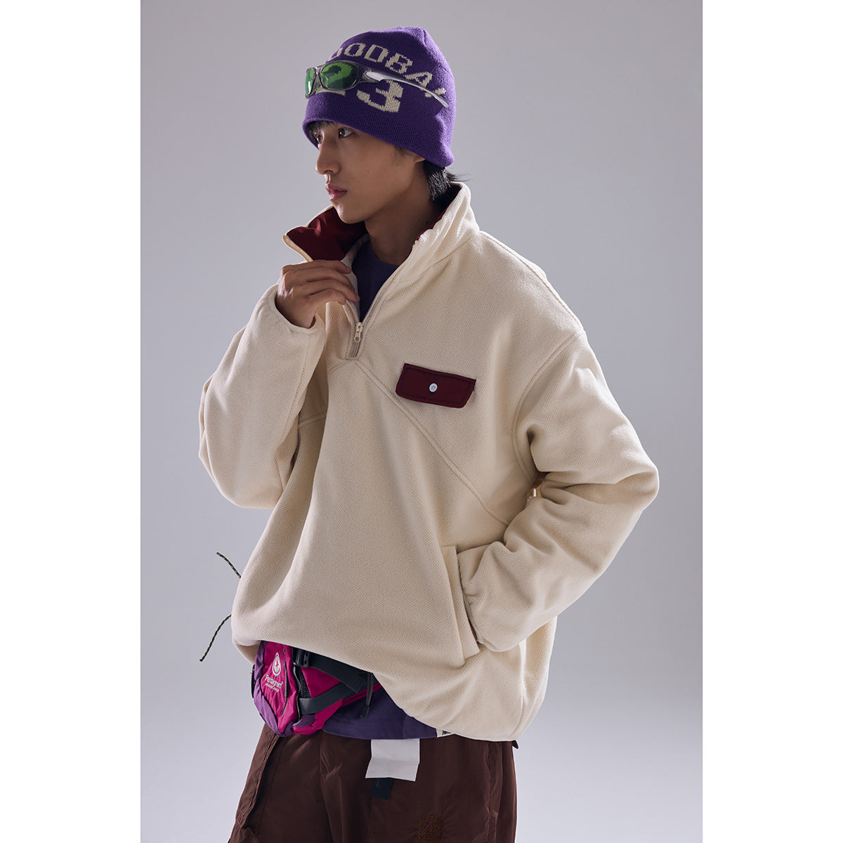 Model wearing an ivory quarter-zip pullover with a burgundy chest pocket and a purple graphic beanie.