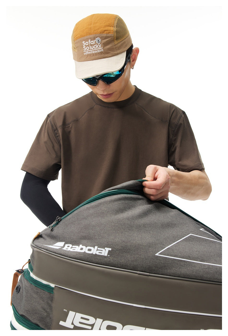 Close view of a model in a brown short-sleeve T-shirt, holding a large gray tennis bag.