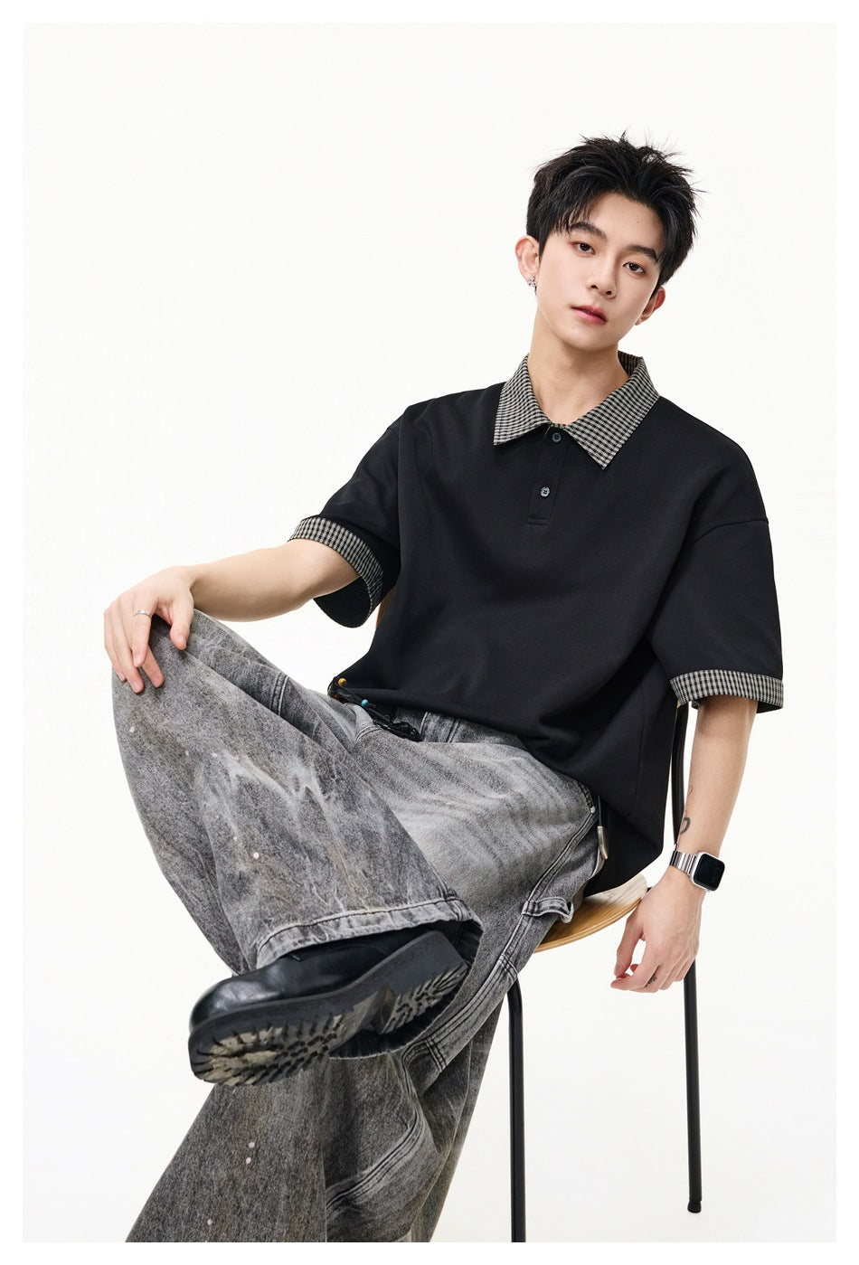 Seated model wearing a loose black polo with checked collar and sleeve cuffs, paired with wide gray jeans.