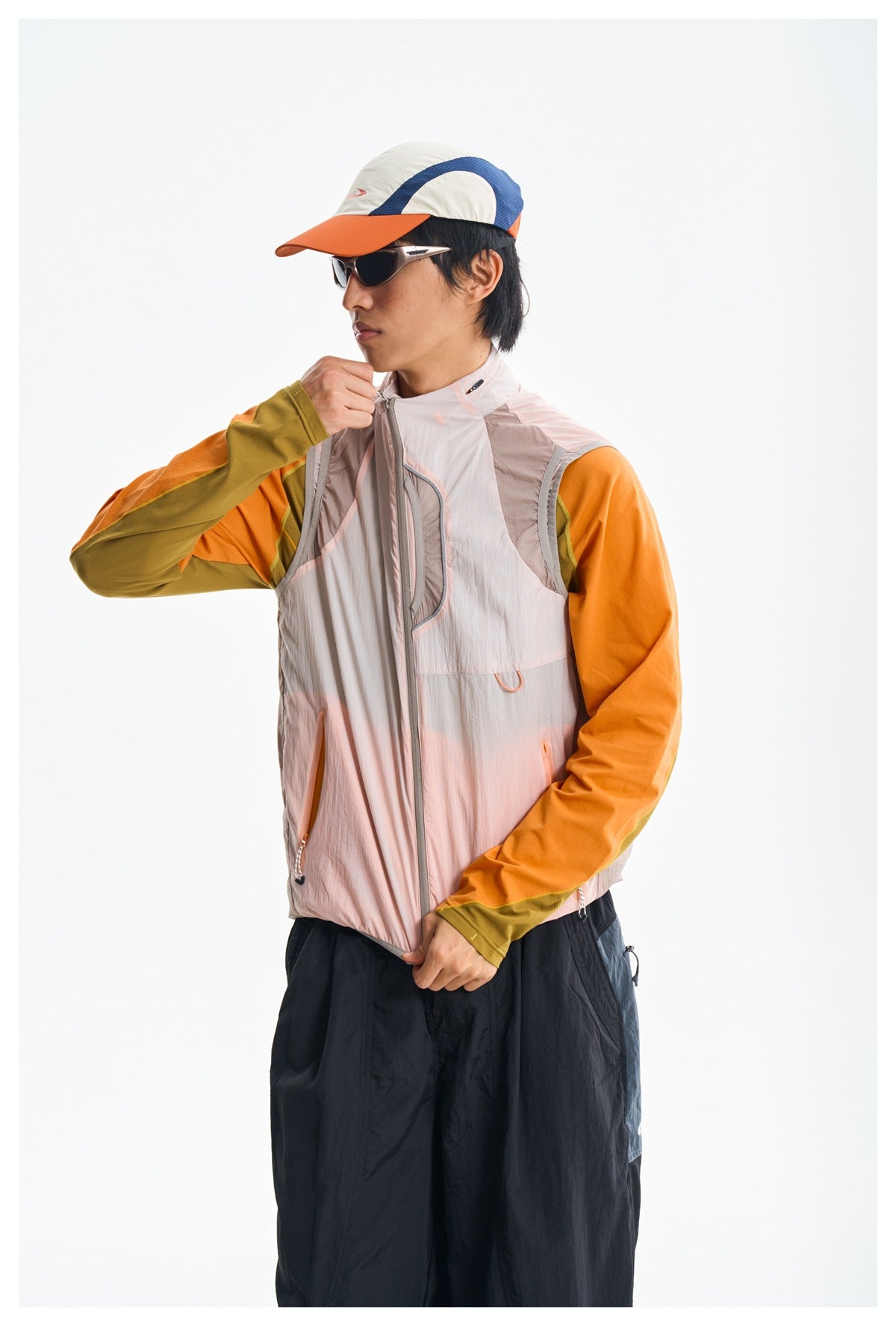 Model adjusting the collar zip of a pale pink vest worn over an orange and olive long-sleeve top.