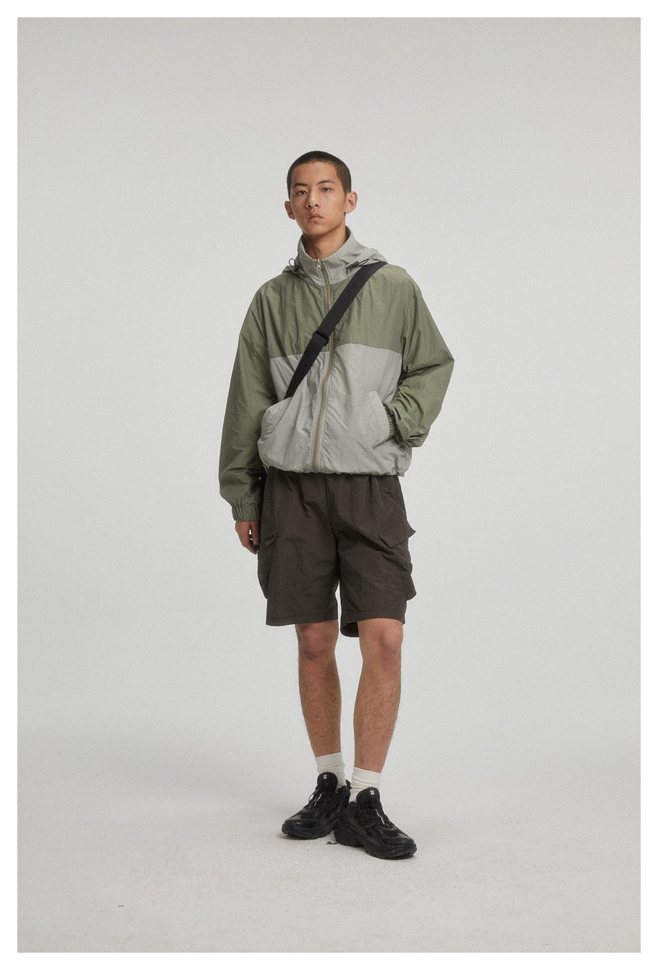Full-length model wearing an olive and light gray color-block hooded jacket, dark cargo shorts, and a black crossbody bag.