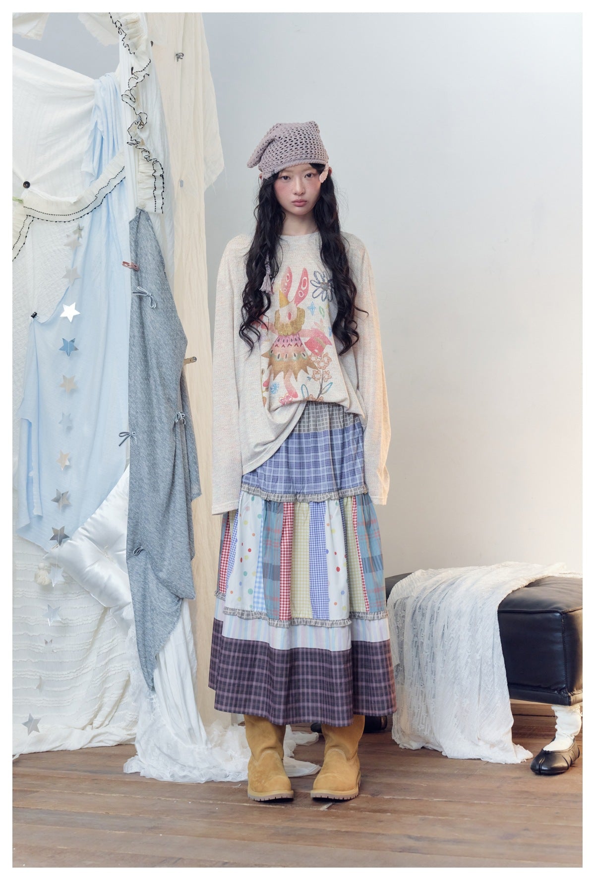 Full-length view of a model in a cream illustrated top and a tiered plaid and multicolor patchwork skirt with tan boots.