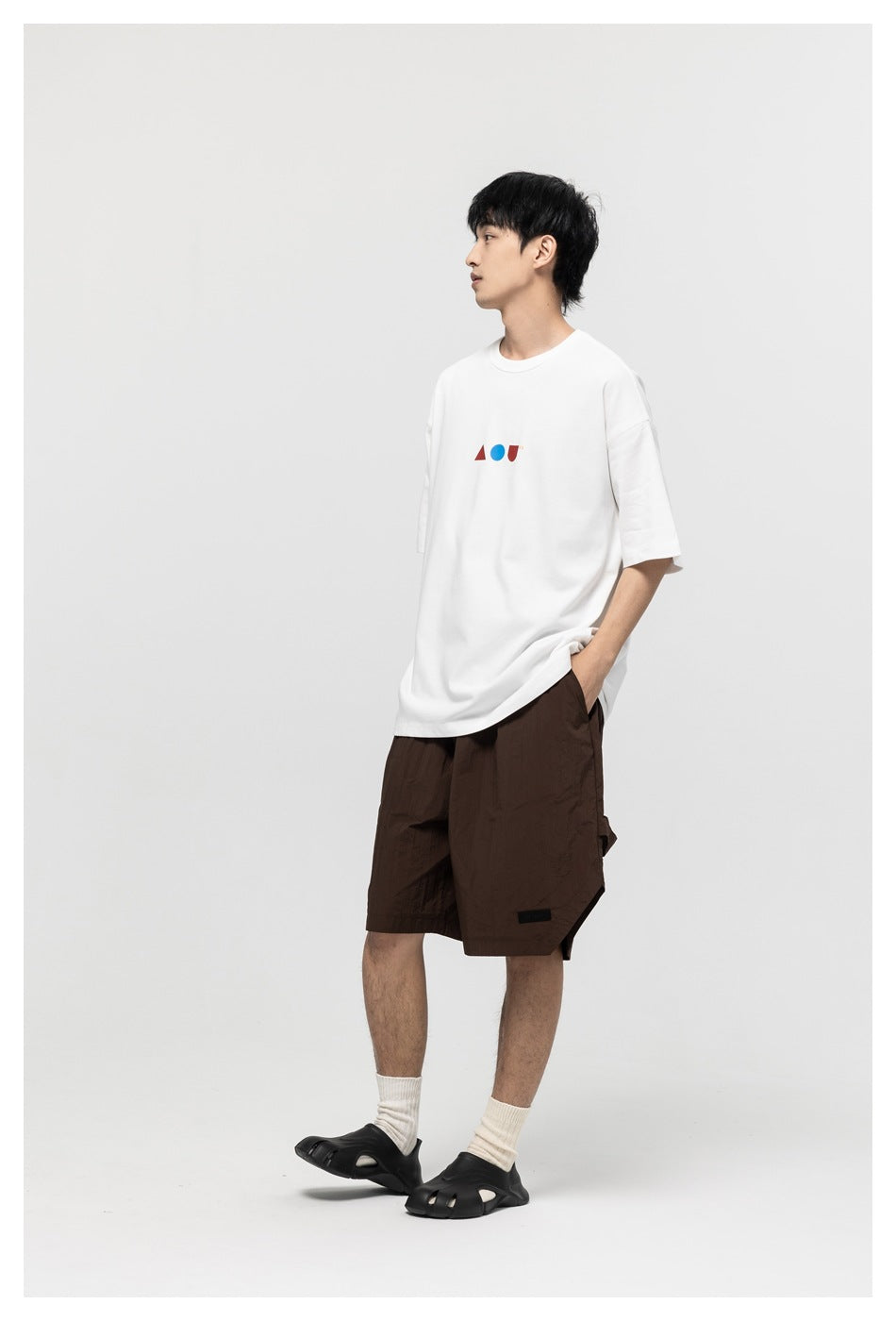 Model in a white oversized T-shirt with small geometric chest graphics and dark brown shorts, shown from a side angle.