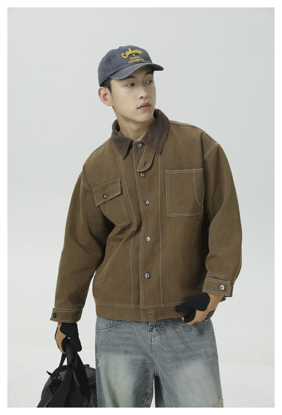 Model wearing a brown work jacket with contrast stitching and flap pockets, paired with a gray cap and light jeans.