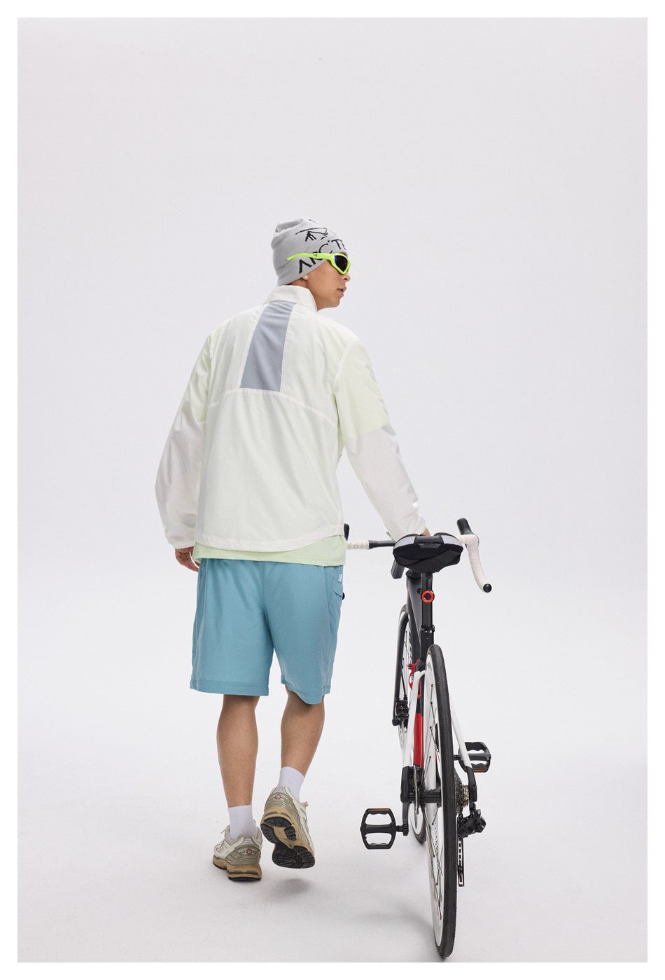Rear view of a man wearing a pale jacket with a gray back panel and blue shorts beside a road bike.