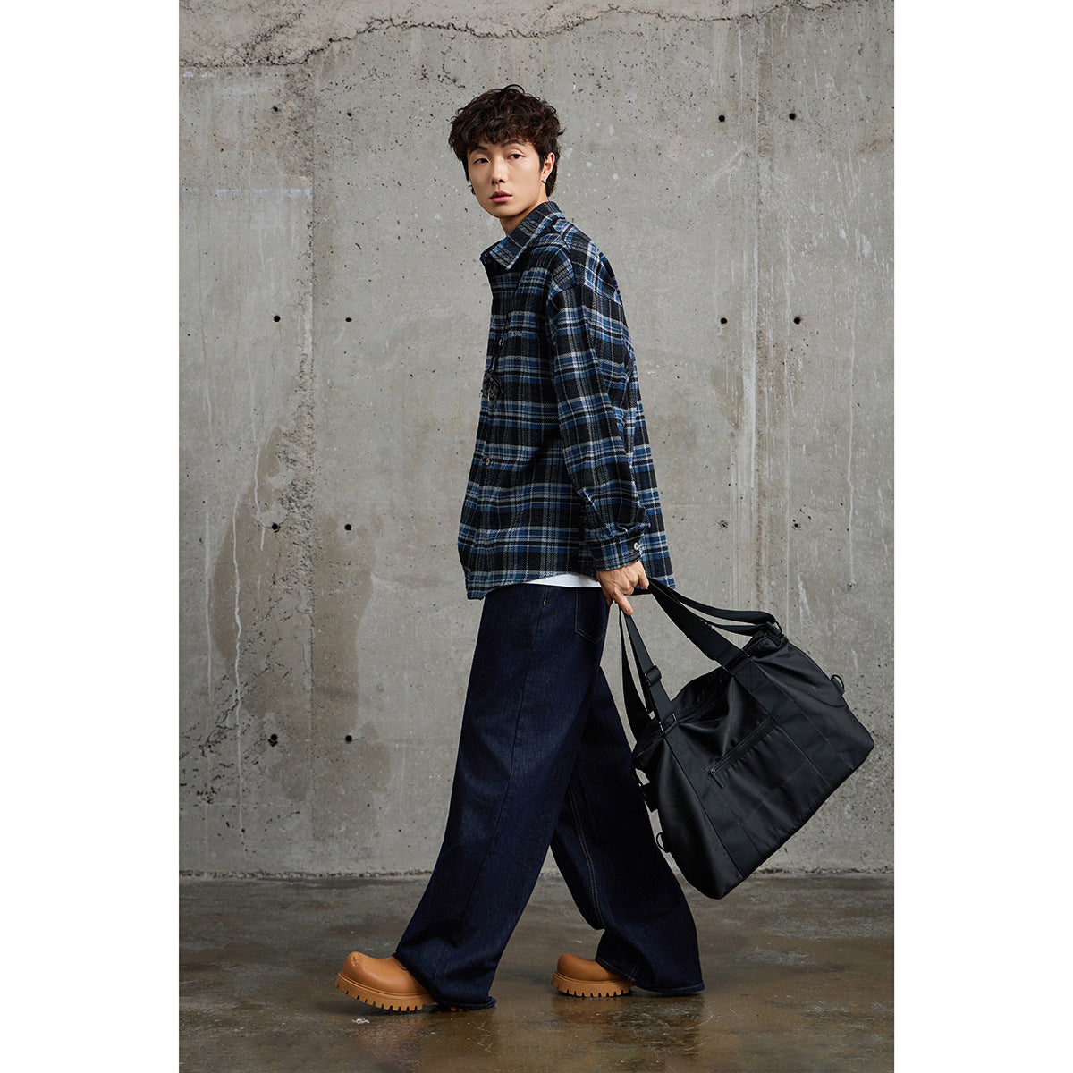 Model wearing a blue-and-black plaid button-up shirt, shown at an angle with a large black bag.