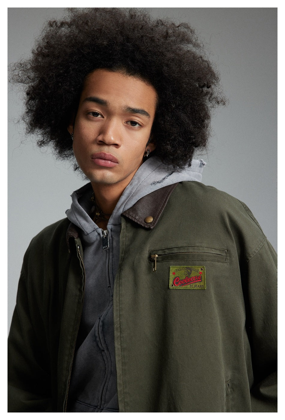 Close view of a model wearing an olive jacket with a brown collar, chest zipper pocket, and embroidered patch.