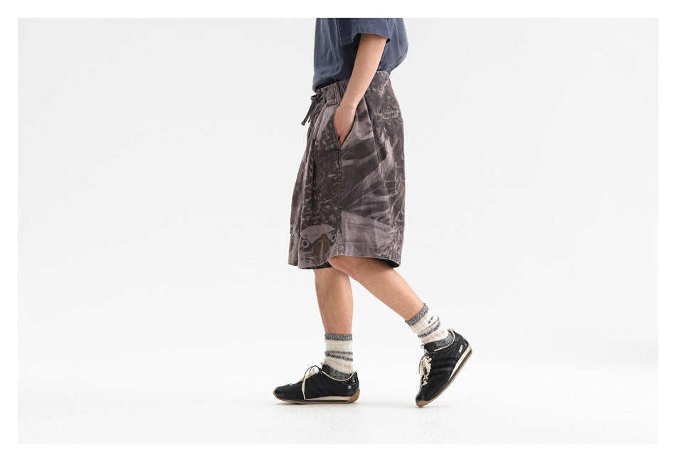 Side view of a model wearing loose charcoal-gray graphic shorts with an elastic waist and wide legs.