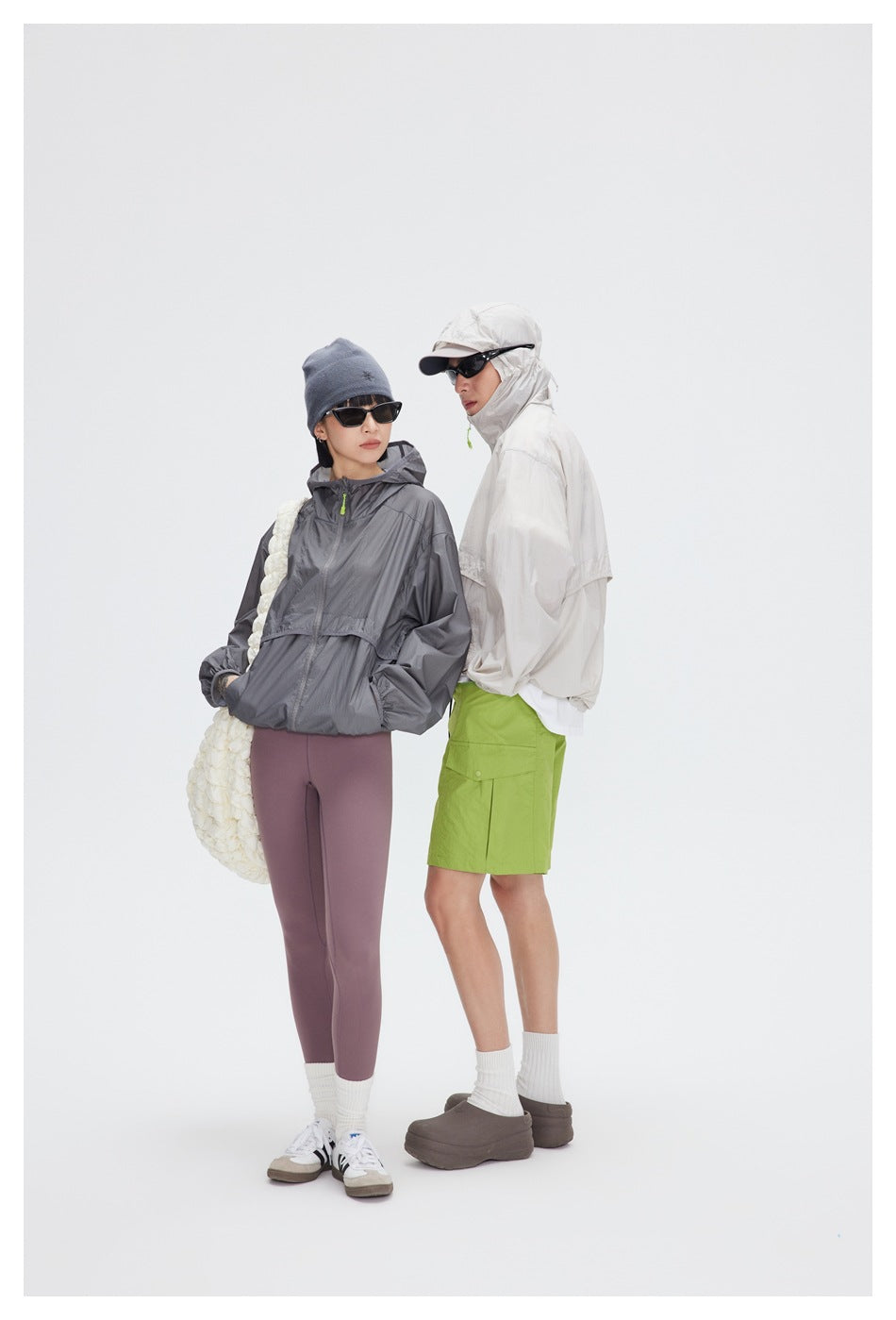 Two models pose in pale beige and charcoal gray hooded jackets; one wears lime-green shorts, the other mauve leggings.