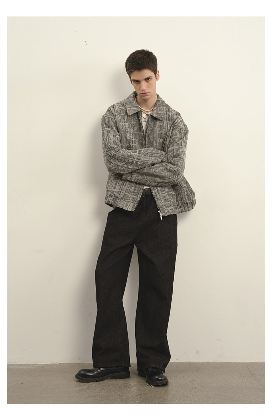 Model leaning on a wall in a gray textured zip jacket with arms crossed, black wide-leg pants and black boots