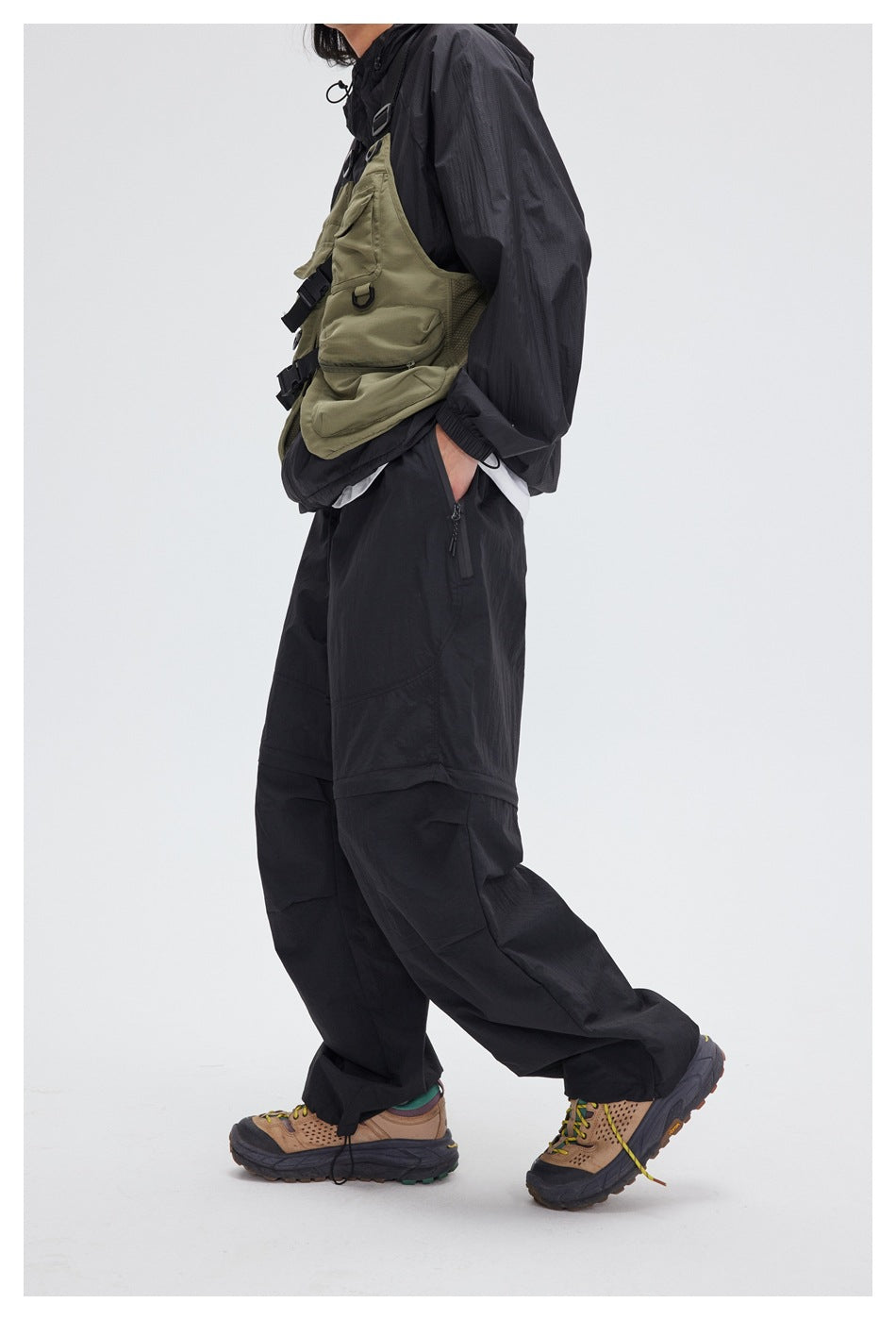Side view of a model wearing loose black panelled pants with a zip pocket and an olive utility vest.