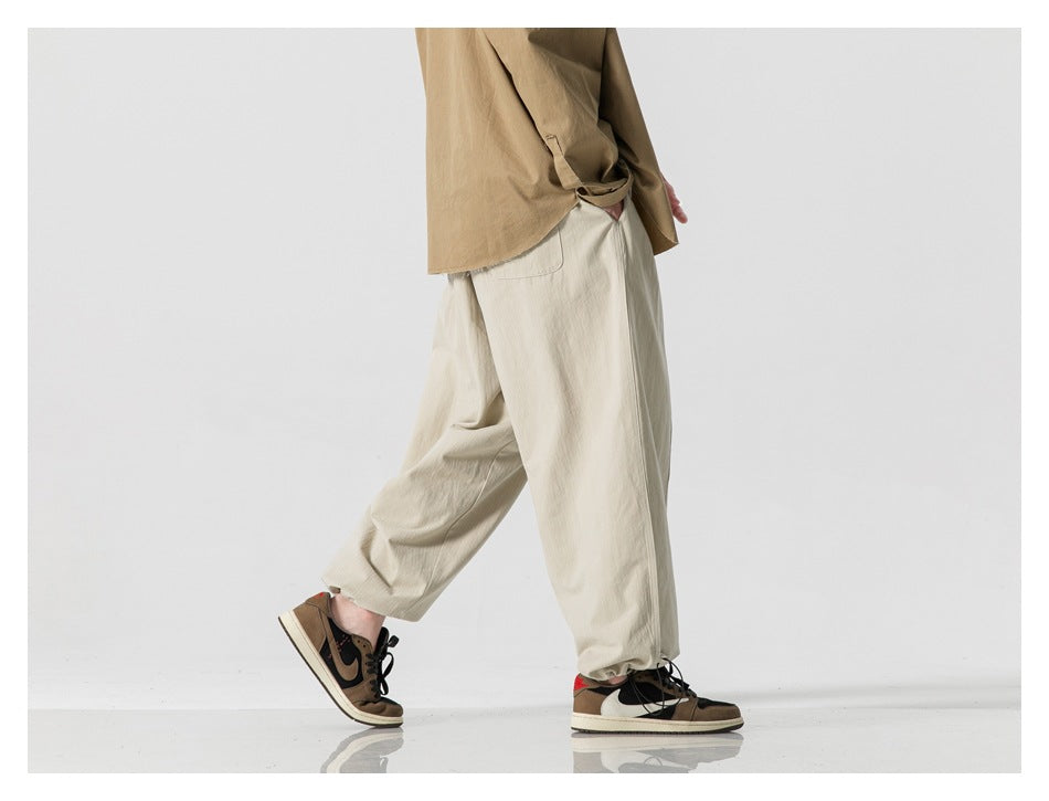 Side view of a model wearing loose light beige trousers with an elastic waist and gathered hems.