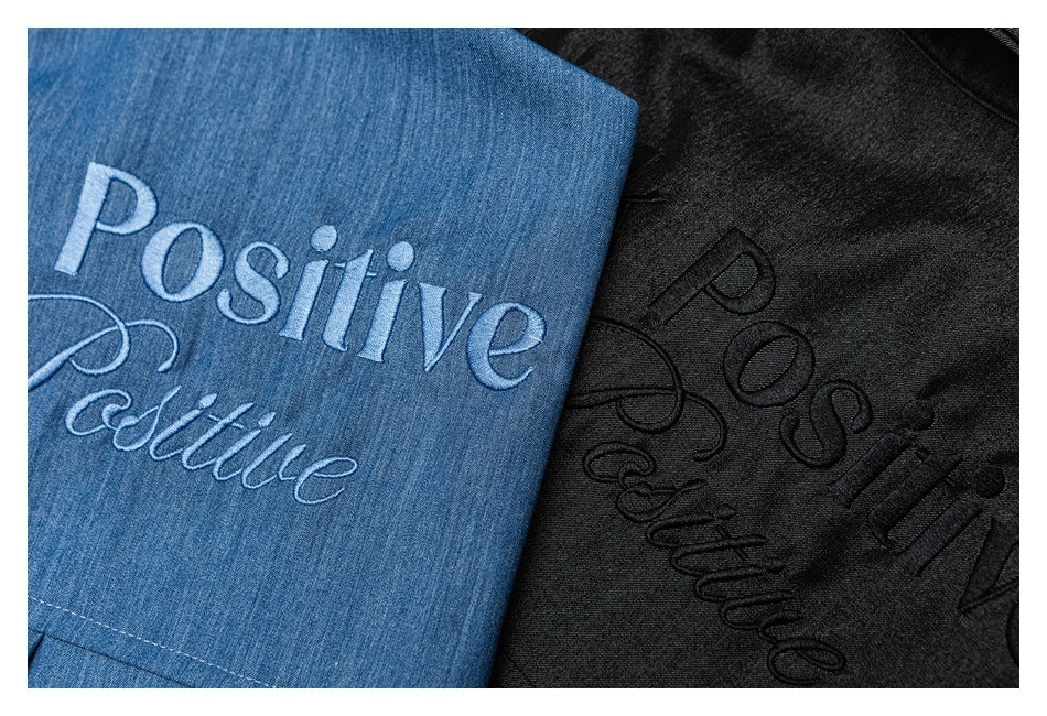 Close-up of blue and black shirts showing contrasting light and dark embroidery reading “Positive.”