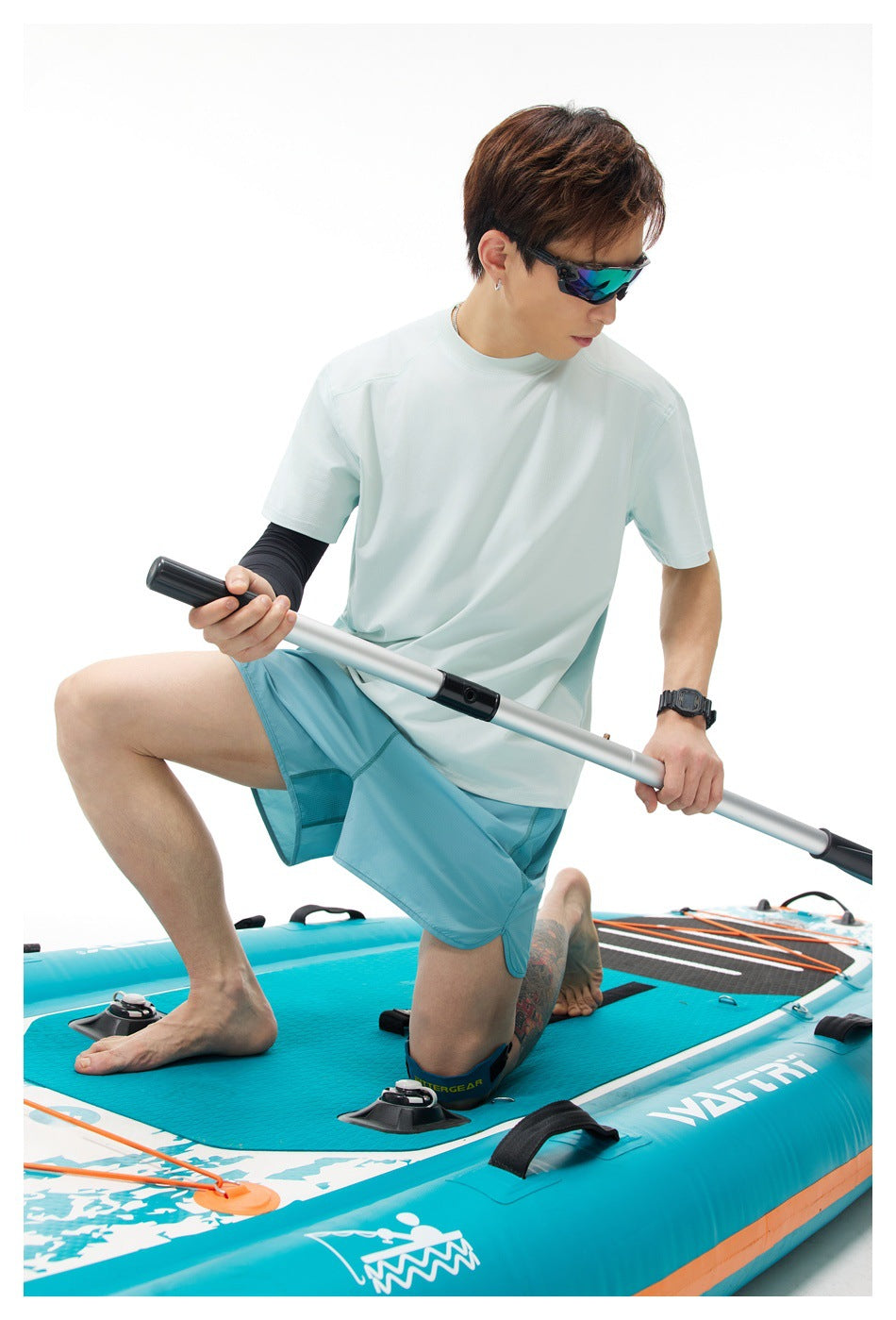 Model wearing a pale mint T-shirt and aqua shorts while kneeling on a turquoise paddleboard with a paddle.