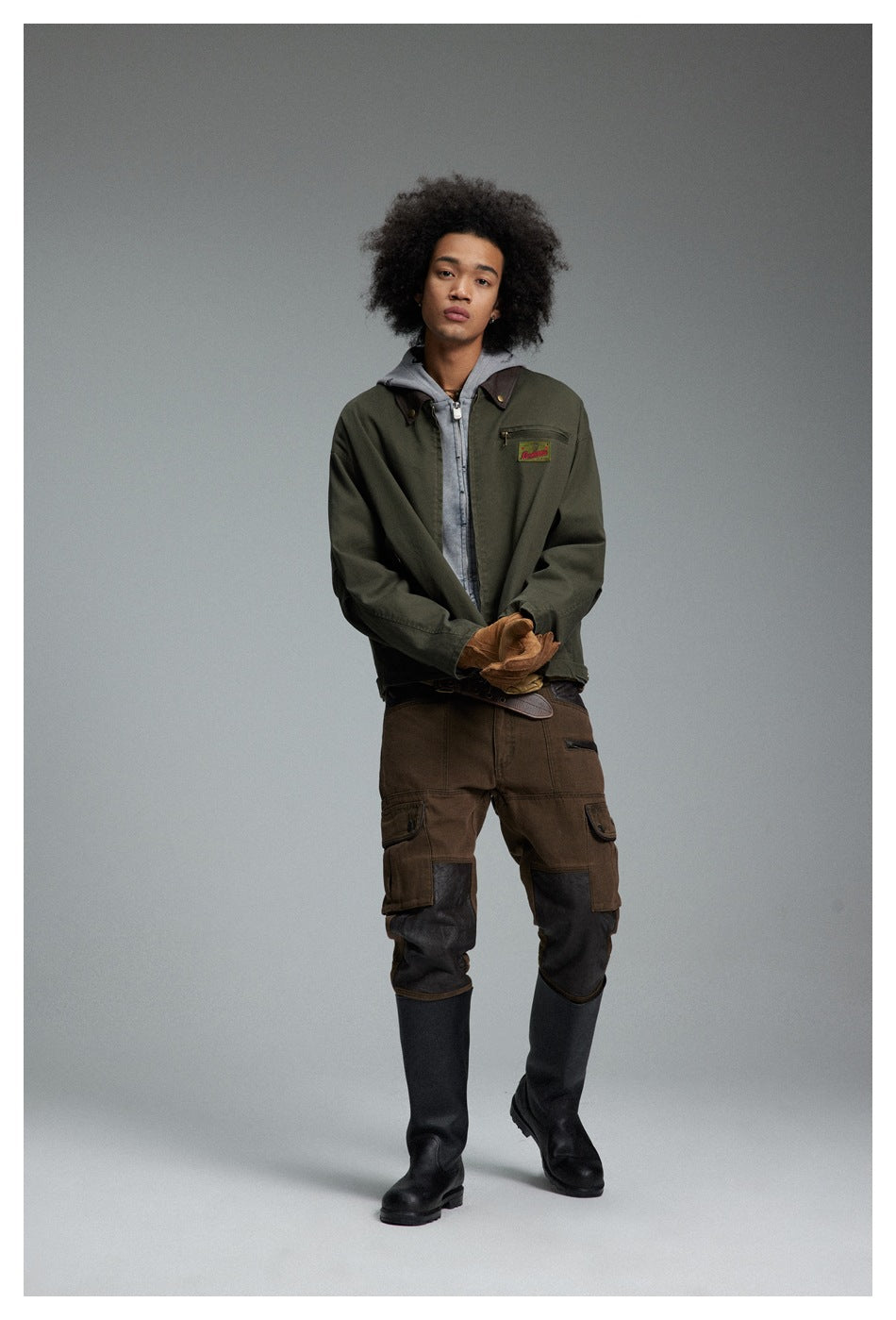 Full-length model wearing an olive work jacket over a gray hoodie with brown cargo pants and black boots.