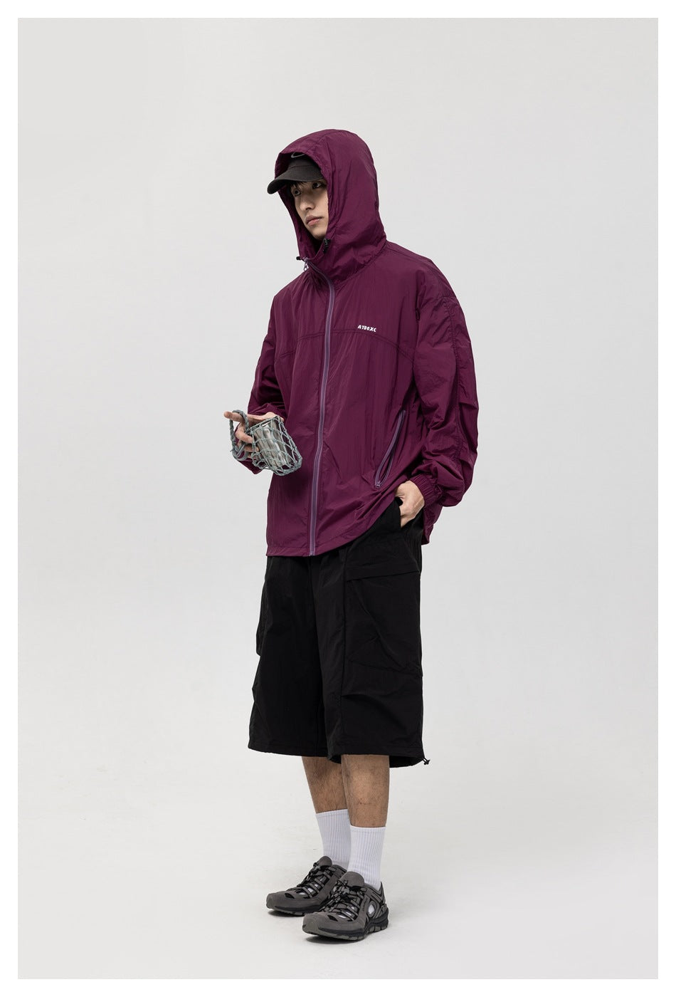 Model in a plum hooded jacket and black shorts, holding a small net bag against a studio backdrop.