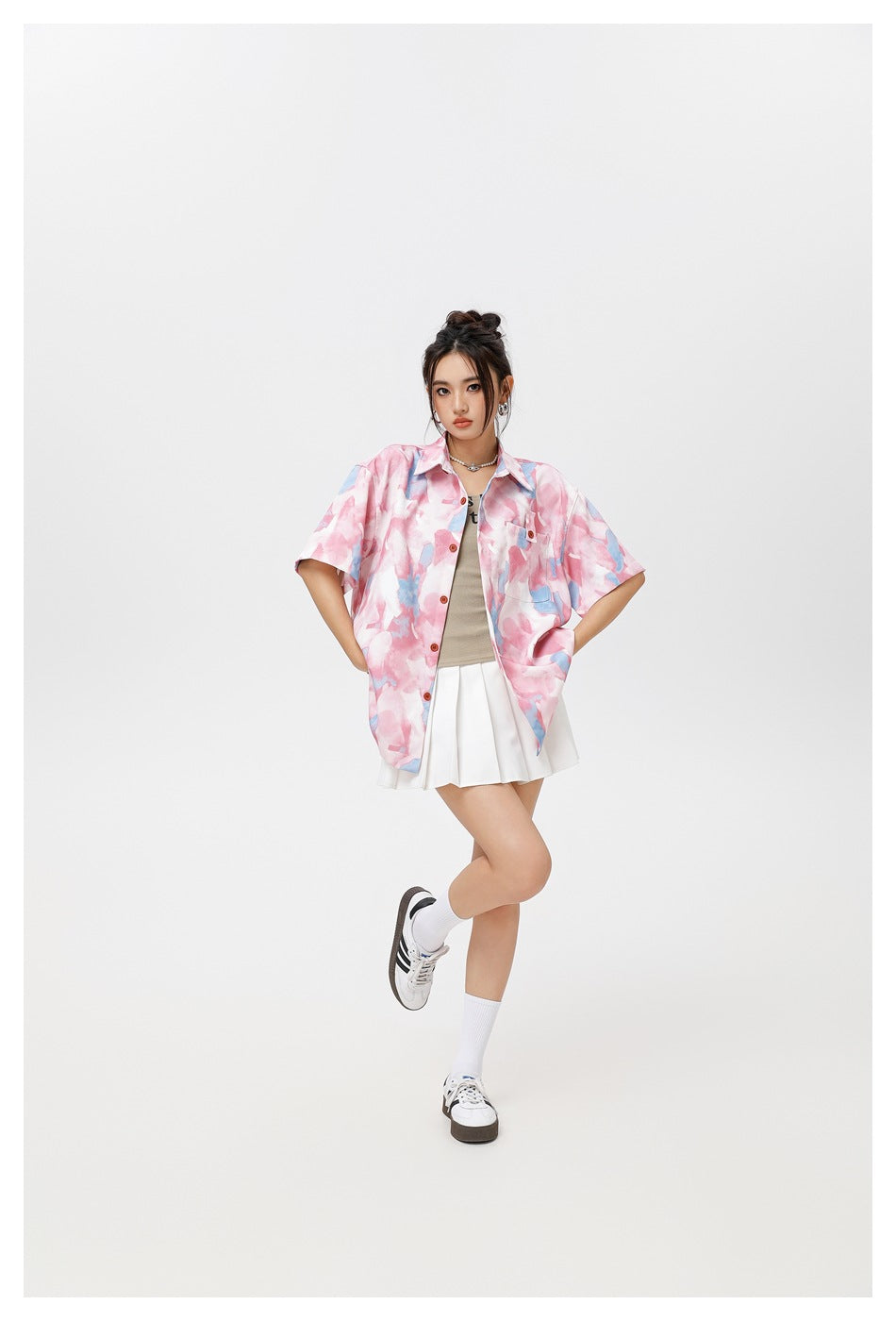 Model posing in a pink and blue abstract-print oversized shirt, white pleated skirt, socks, and sneakers.