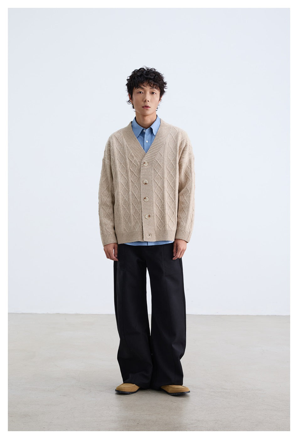 Model wearing a beige diamond-knit button cardigan over a blue shirt with black wide-leg trousers, front view.