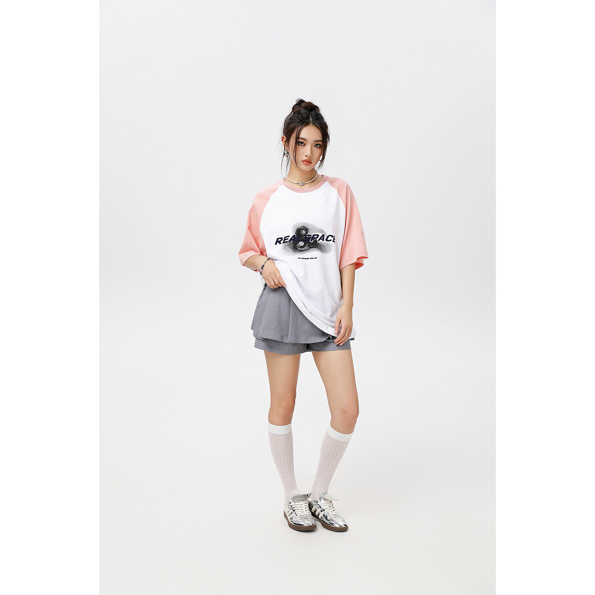 Model wearing an oversized white raglan T-shirt with pale pink sleeves and a black chest graphic, paired with gray shorts.