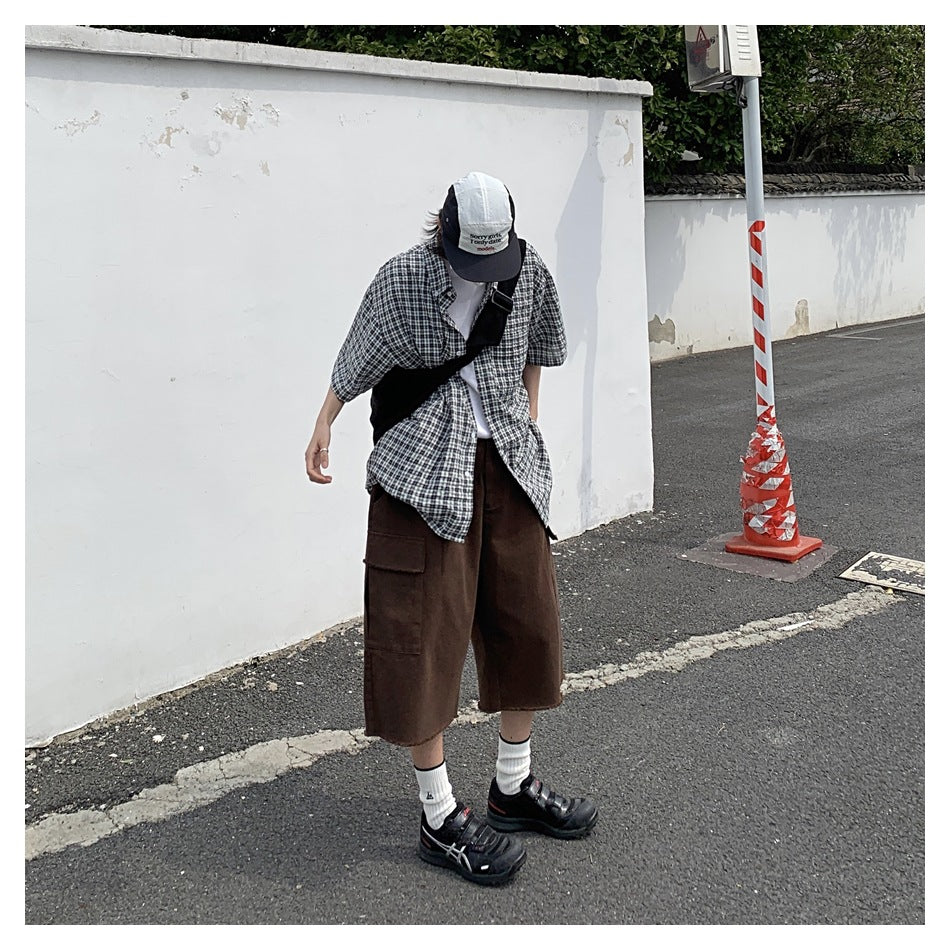 A person stands outdoors wearing a dark plaid short-sleeve button-up shirt, brown shorts, and a cap.