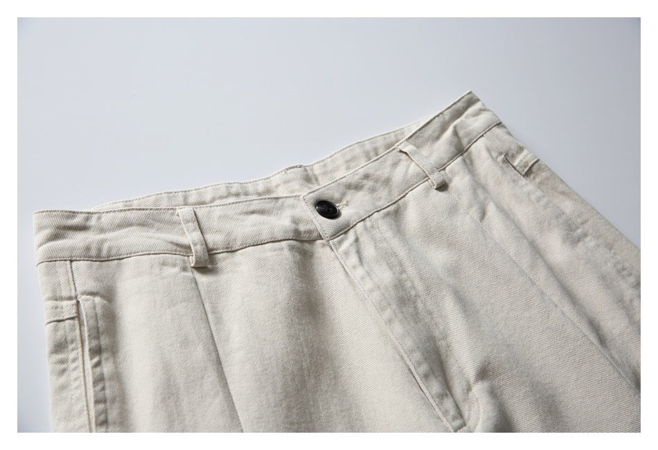 Close-up of the front waistband of light beige trousers, showing a black button, belt loops and pleats.
