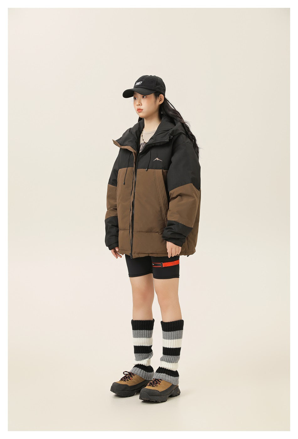 Model wearing a black and brown color-block puffer jacket with a cap, black shorts and striped leg warmers