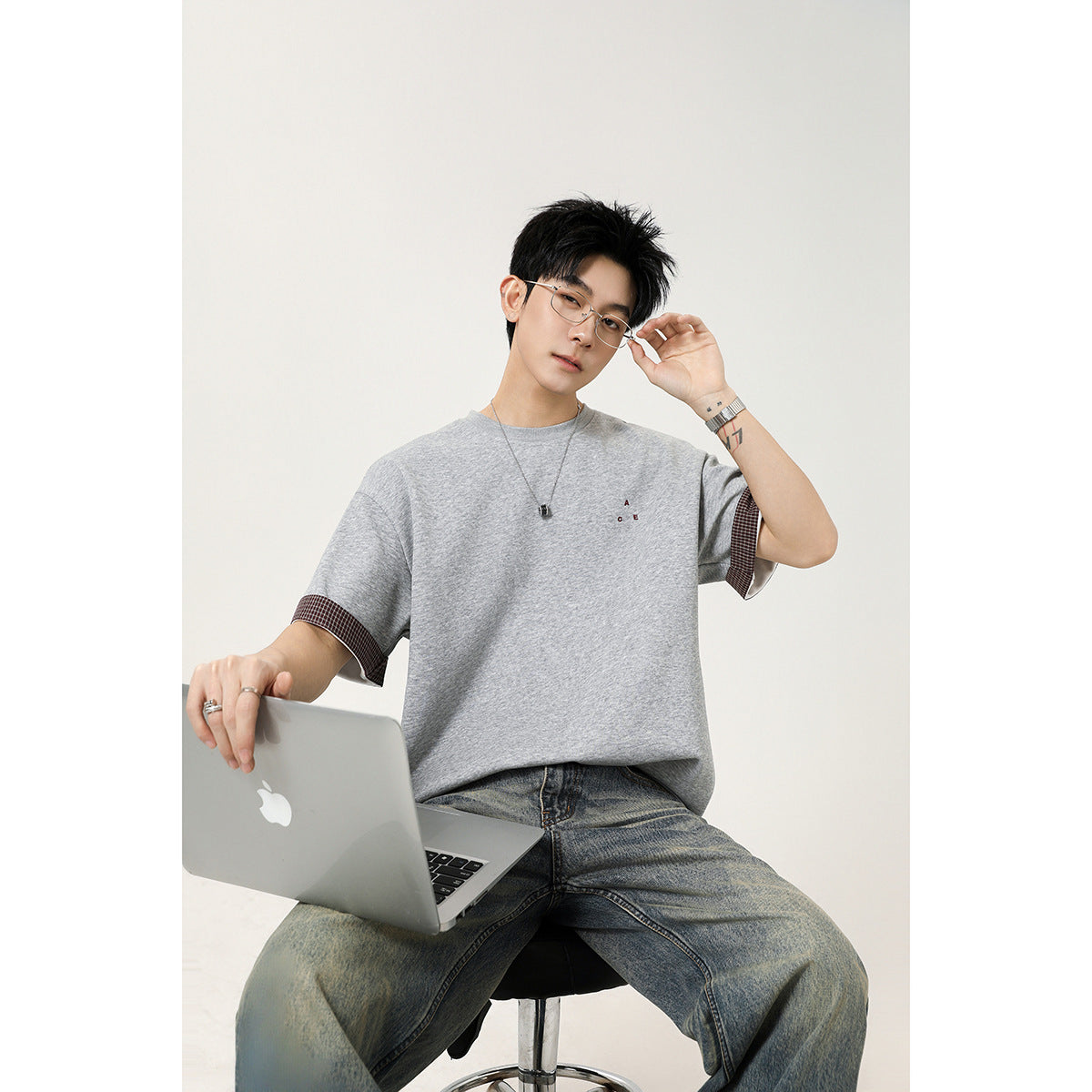 A seated man wears an oversized light gray T-shirt with dark checked sleeve cuffs and a small chest design.