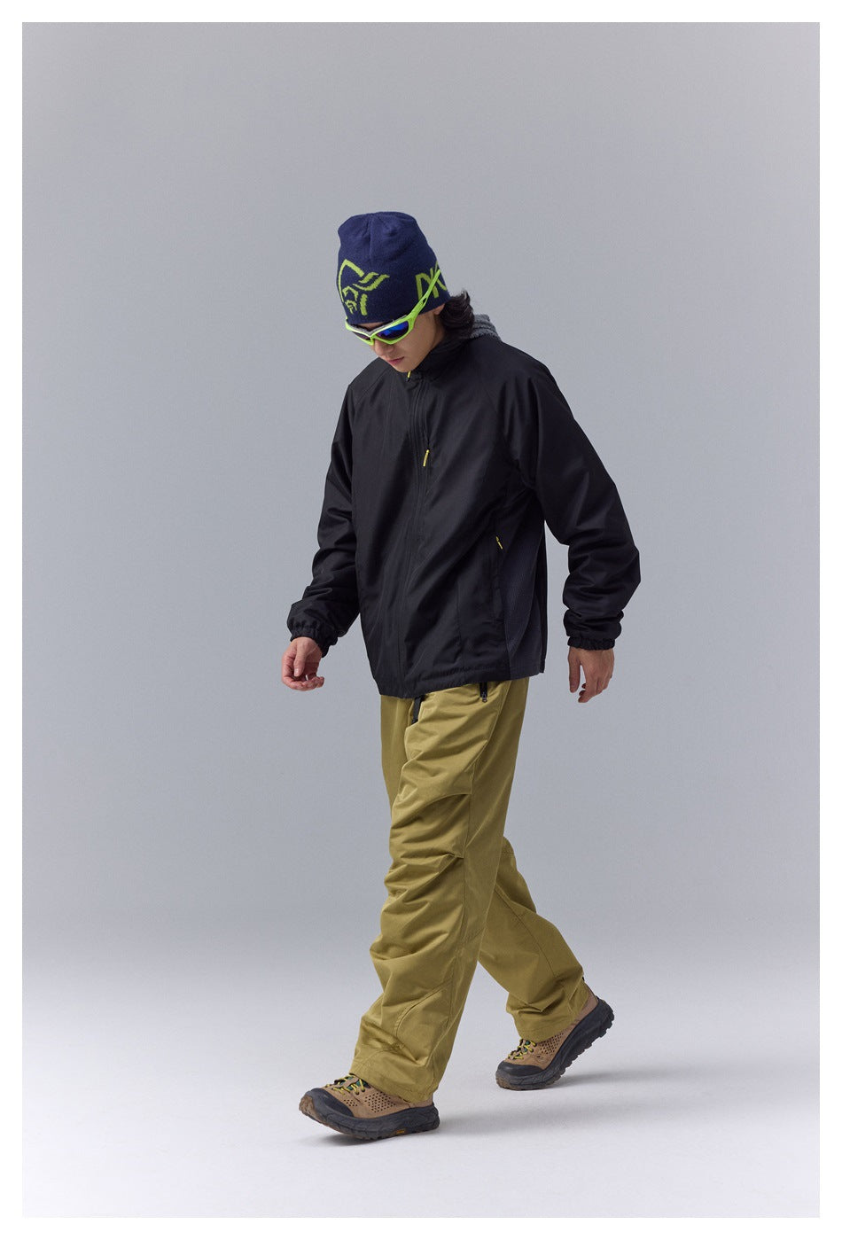 Model walking in a black zip jacket with yellow accents and olive trousers.