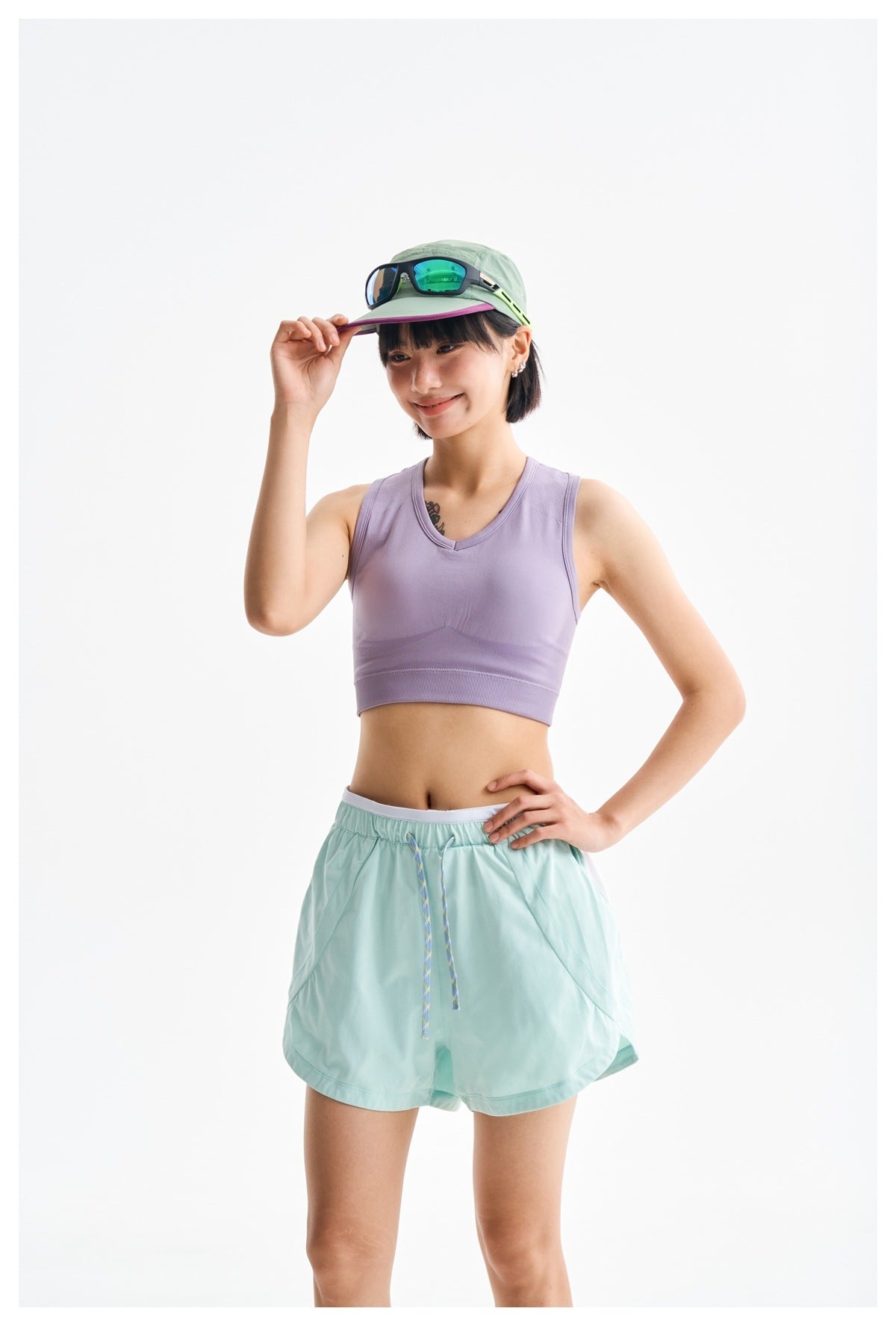 Model wearing a lavender sleeveless sports top, mint running shorts, and a pale green cap with sunglasses.