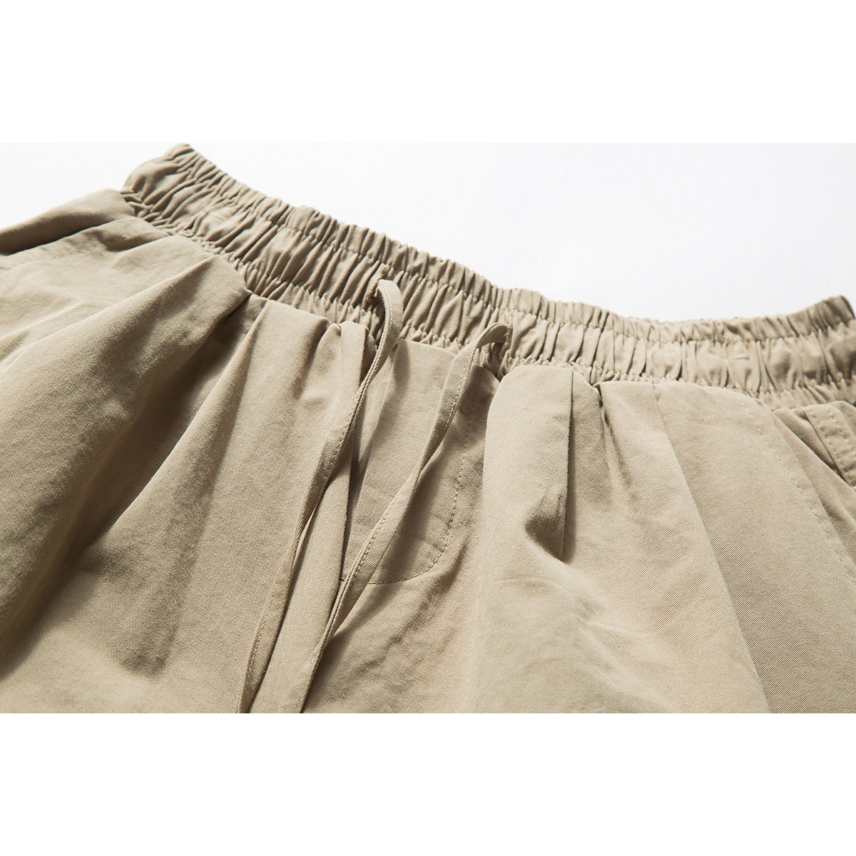 Close-up of tan shorts showing the gathered elastic waistband, drawstring, front pleats and pocket opening.