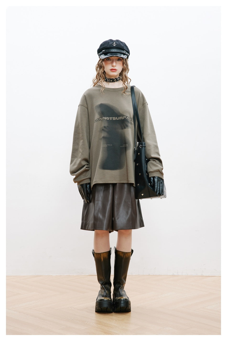 Model wearing an oversized gray-green graphic sweatshirt with leather-look shorts, boots, cap, gloves and a black bag
