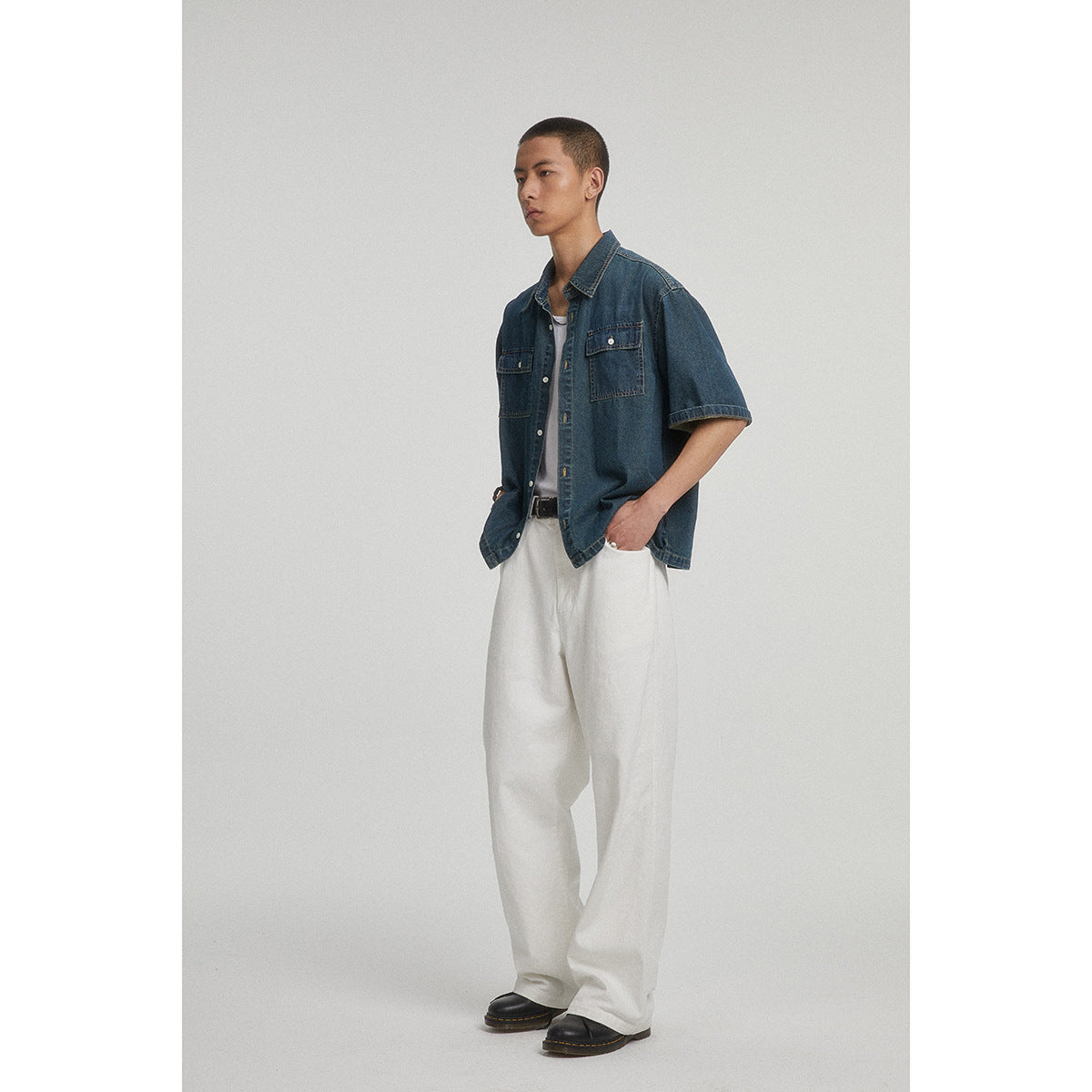 Model wearing a dark blue short-sleeve denim shirt with two chest pockets over a white tee, with loose white trousers.