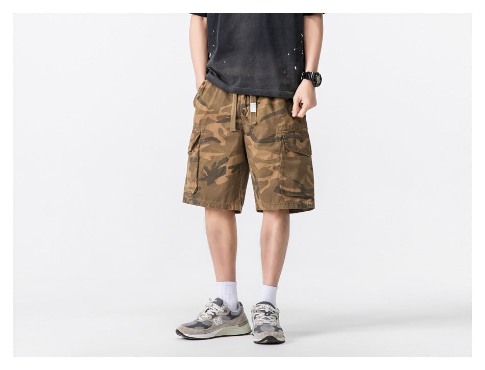 Person wearing khaki camouflage cargo shorts with a dark tee and gray sneakers, front view, cropped at the chest