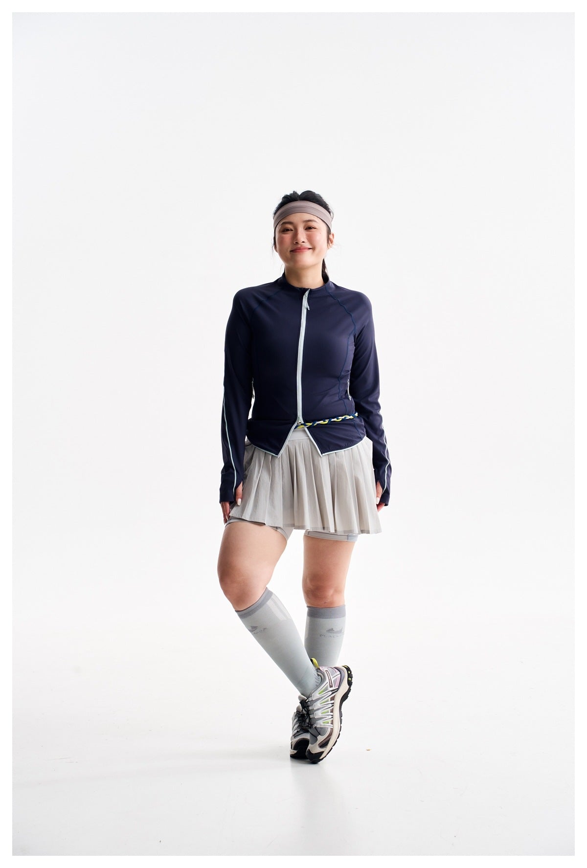 A woman models a navy zip-front athletic jacket with a light gray pleated skort and fitted shorts.