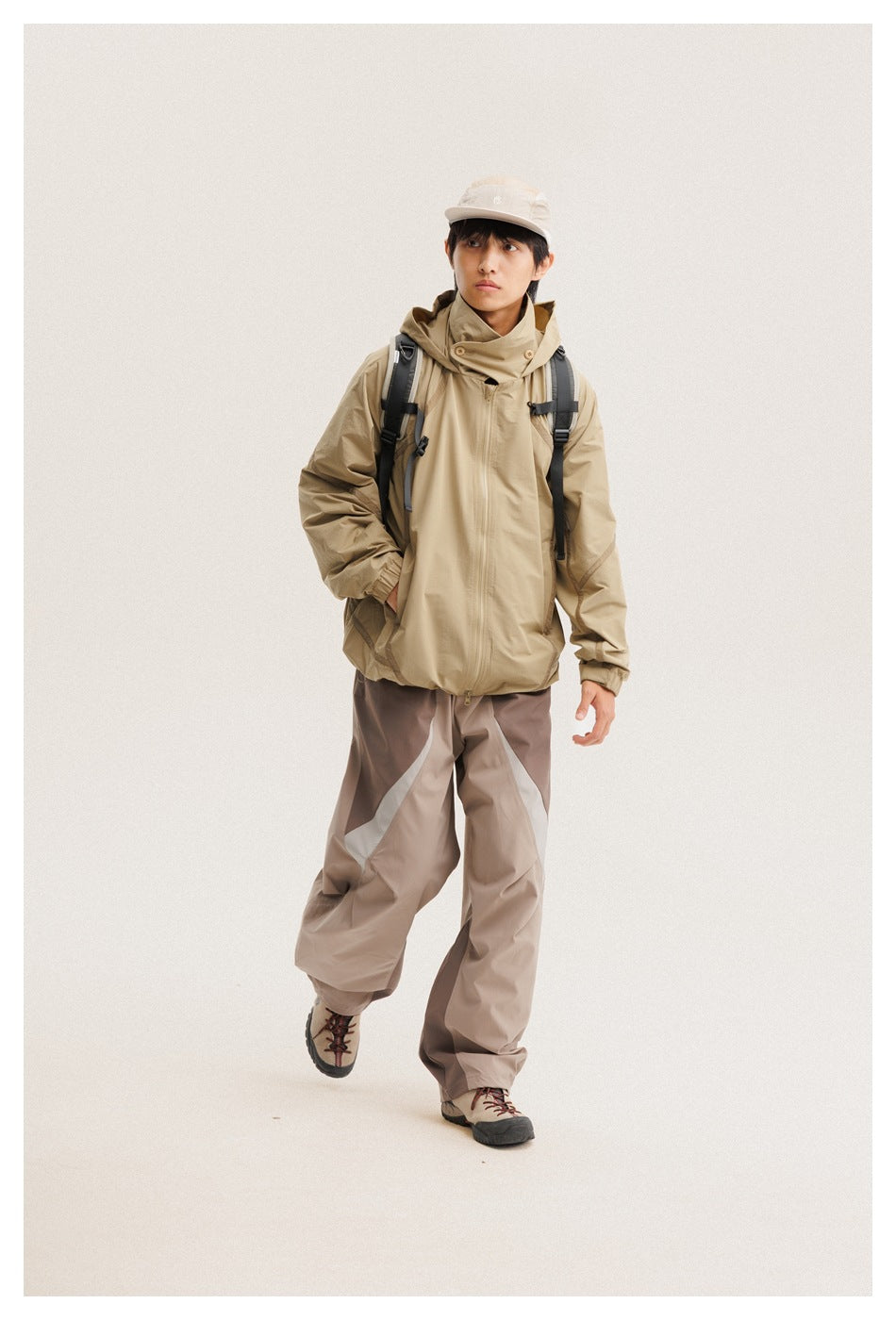 Model wearing a tan hooded zip jacket, taupe color-block pants, a cream cap, and hiking boots.