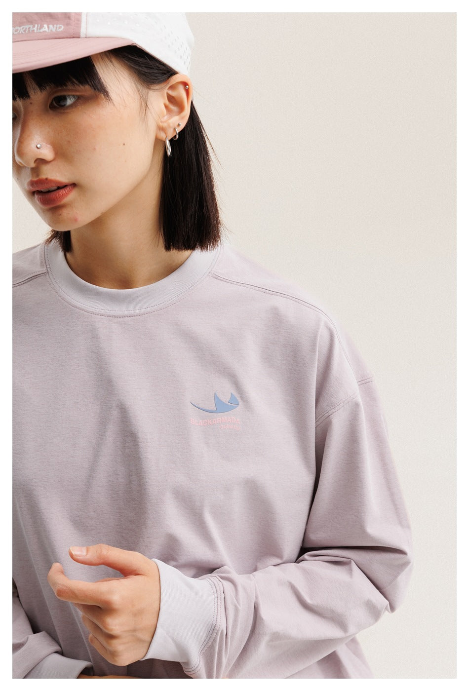 Close-up of a model wearing a pale lavender long-sleeve crewneck shirt with a small chest graphic and a white-and-pink cap.