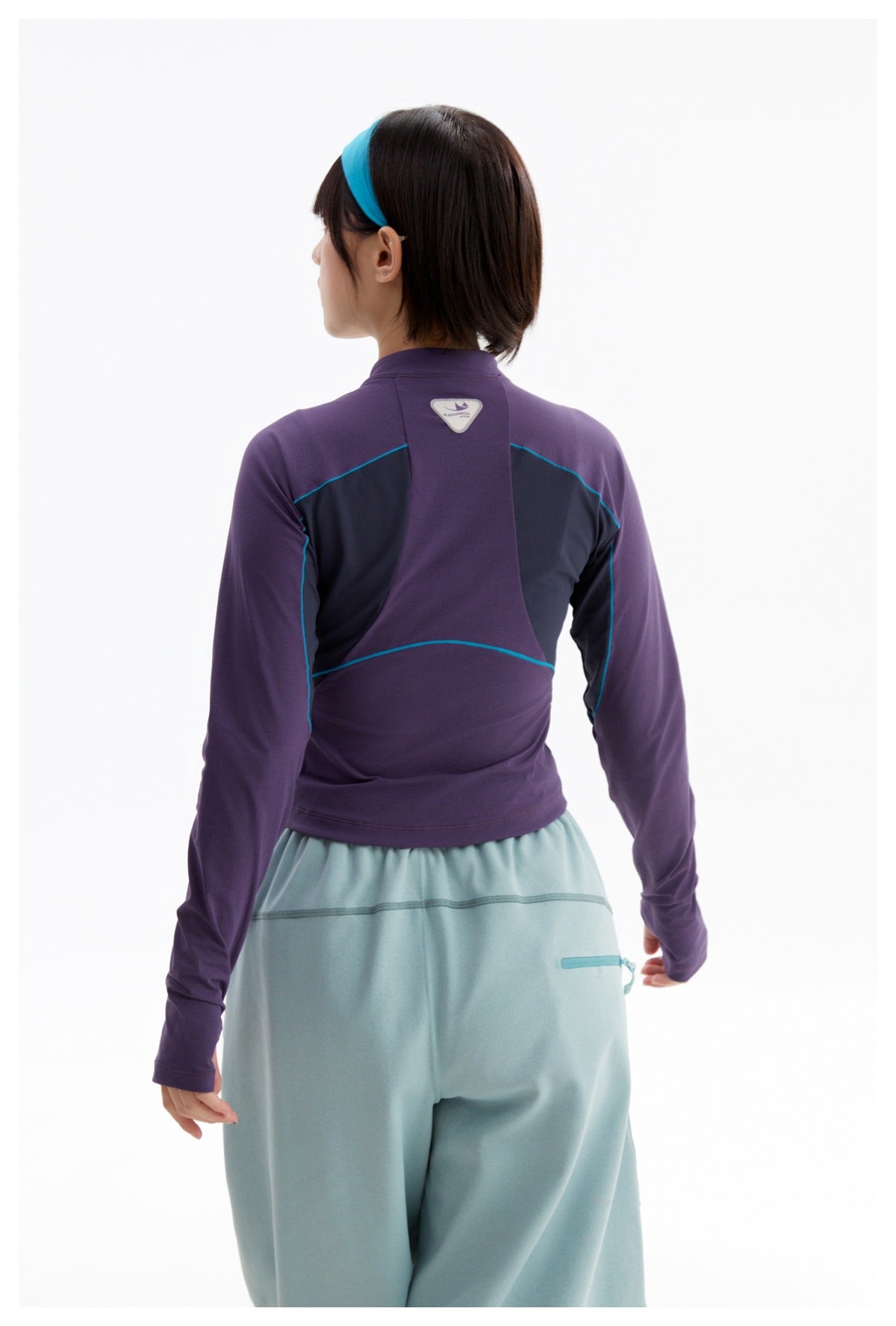 Back view of a model wearing a purple long-sleeve top with dark panels and teal piping, plus pale blue trousers.