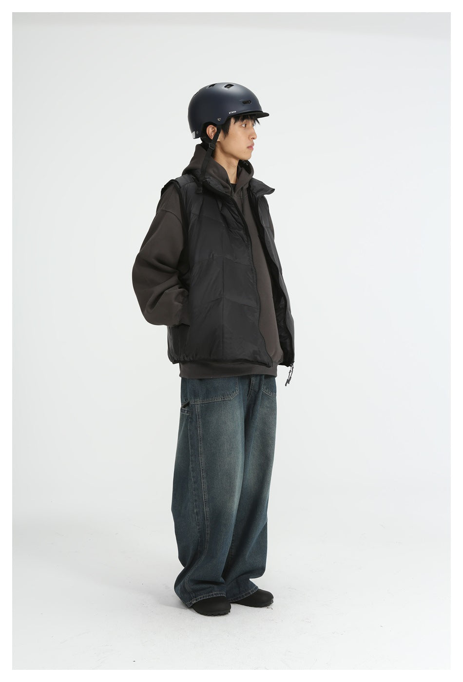 Model wearing a black sleeveless puffer vest over a dark hoodie with loose blue jeans and a helmet.