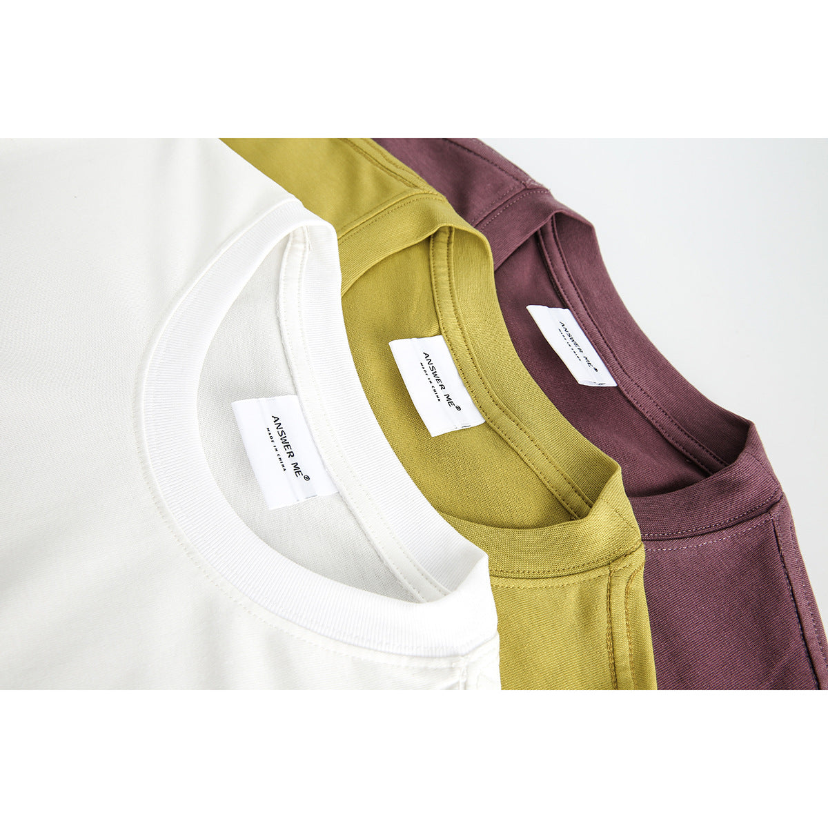 Close-up of three T-shirt necklines in white, mustard yellow, and burgundy, with ribbed collars and sewn labels.