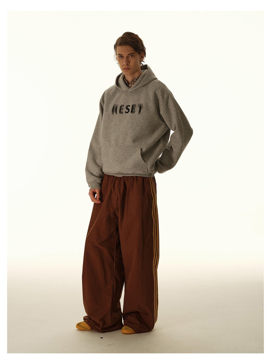 Model wearing a gray printed hoodie and loose brown track pants with side stripes, in a three-quarter pose.