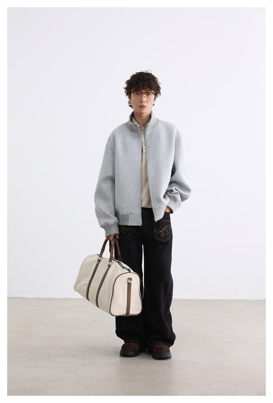 Full-length model wearing a light gray zip jacket and black jeans, carrying a cream duffel bag.